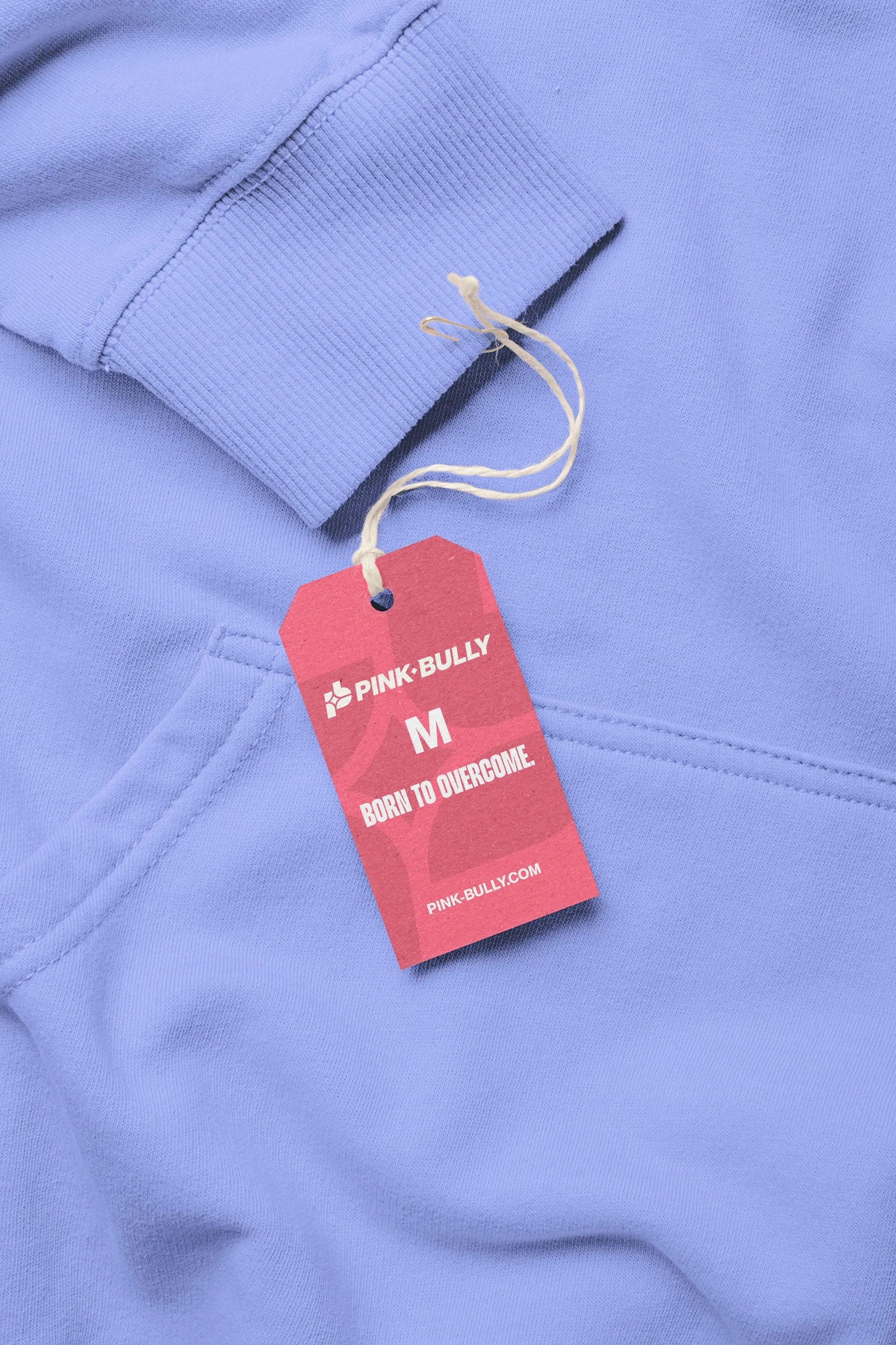 Close-up of a light purple hoodie with a red clothing tag that reads 'Pink Bully, M, Born to Overcome' attached to the hoodie.