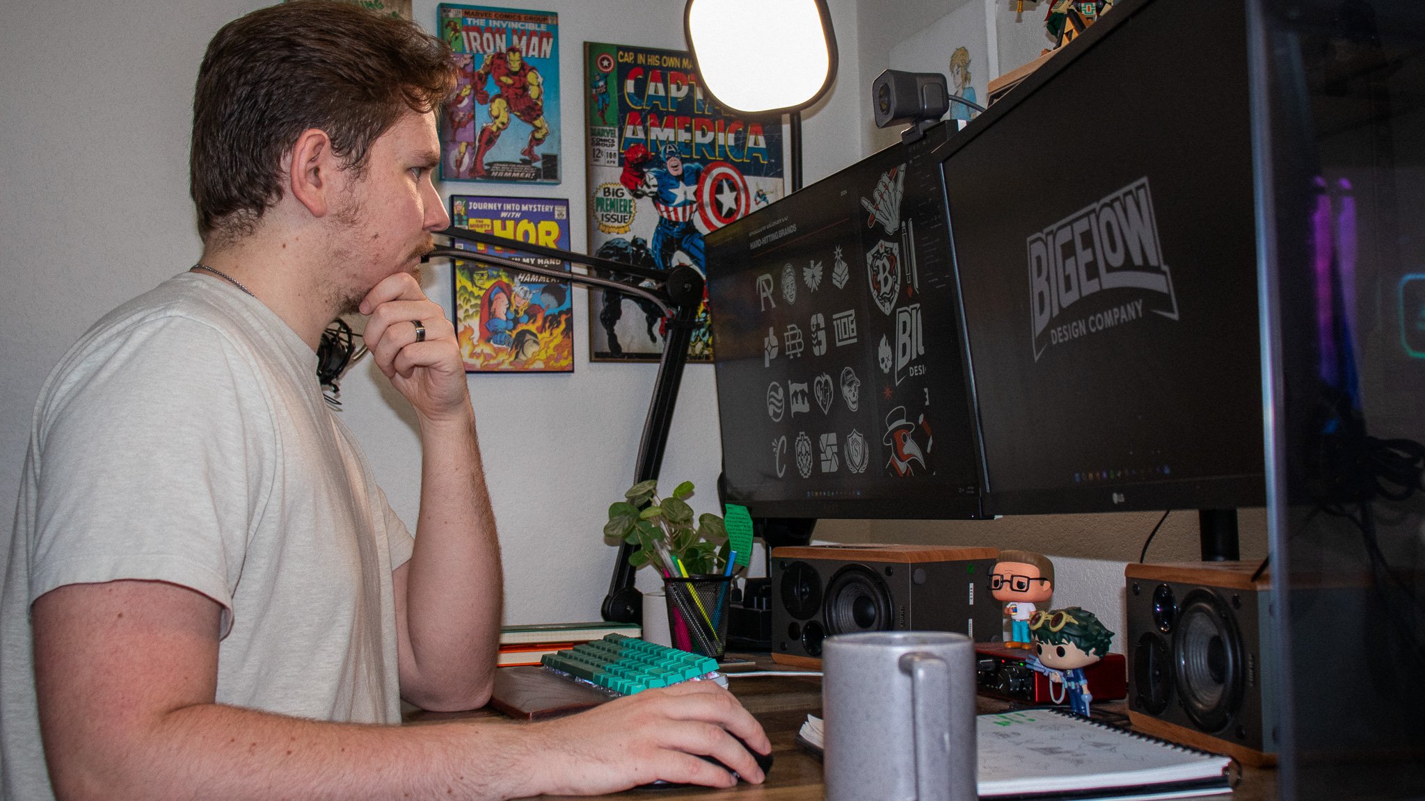 A man with curly brown hair and a beard, wearing a beige T-shirt, sitting at a desk with a computer monitor, keyboard, and speakers, with Marvel comic book posters on the wall behind him.
