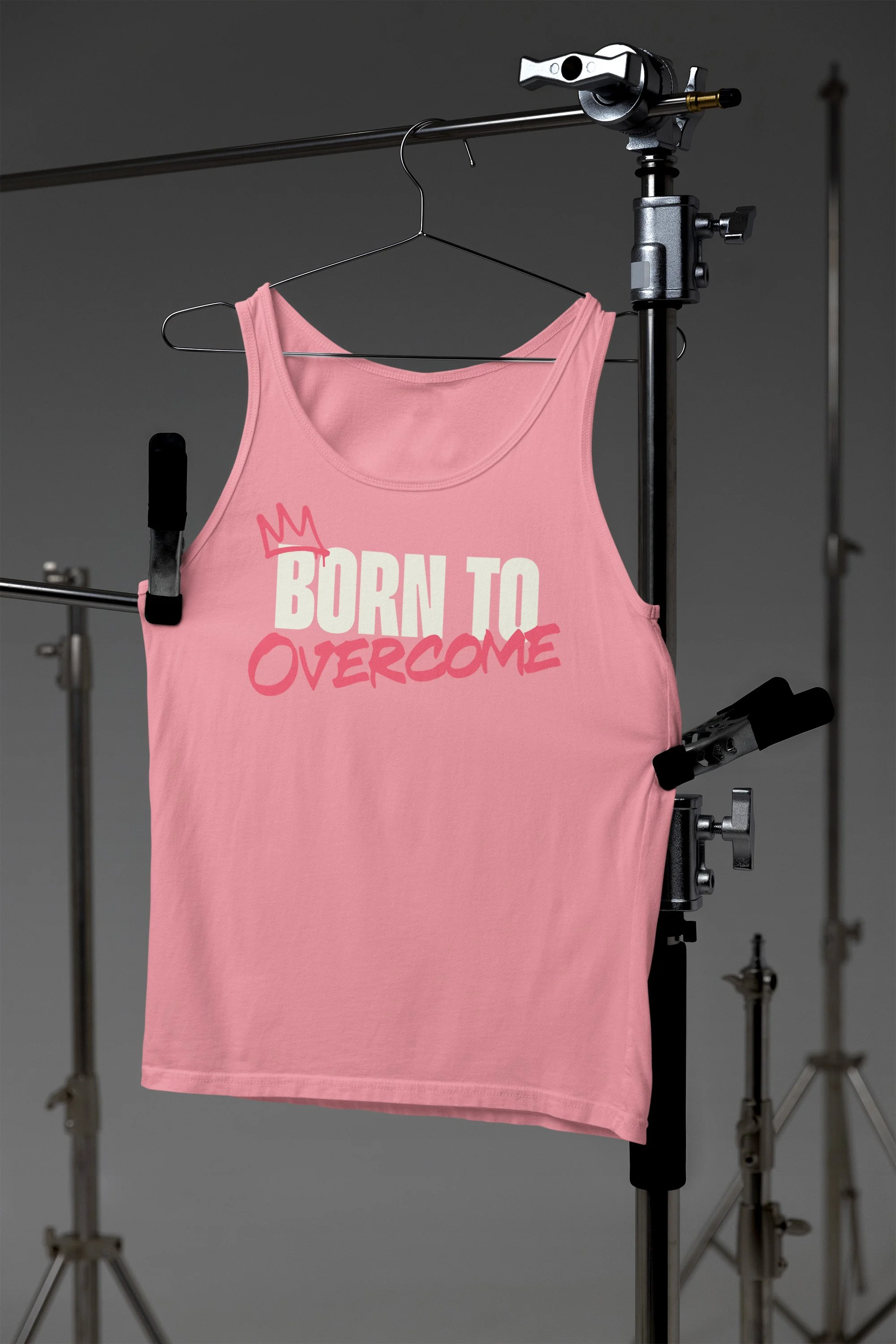 A pink tank top with the words 'BORN TO OVERCOME' printed in white and pink, hanging on a hanger on a clothing rack in a photography studio setting.