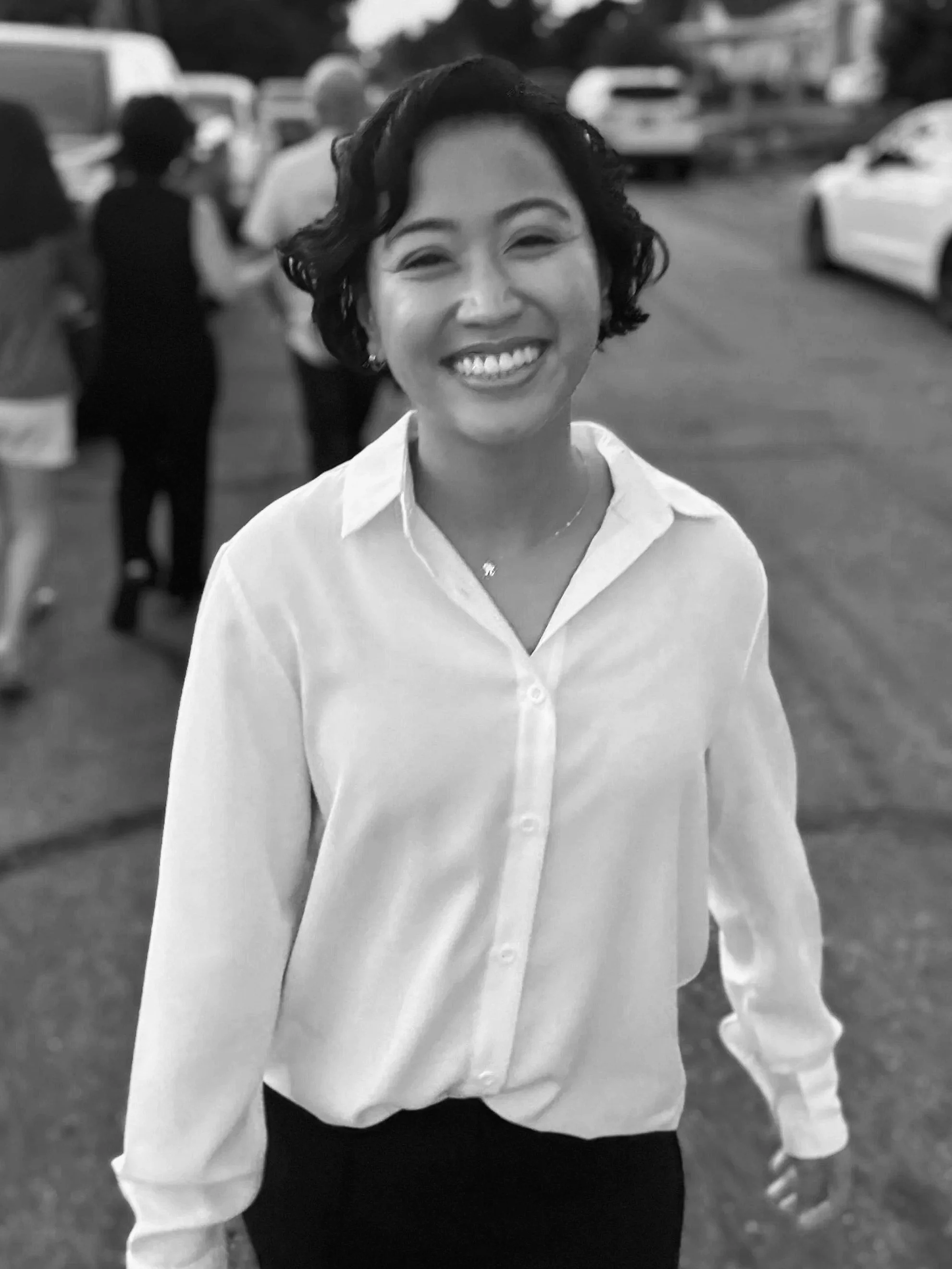 Helen Bass - A smiling woman in a white shirt walking outdoors, with cars and people in the background.