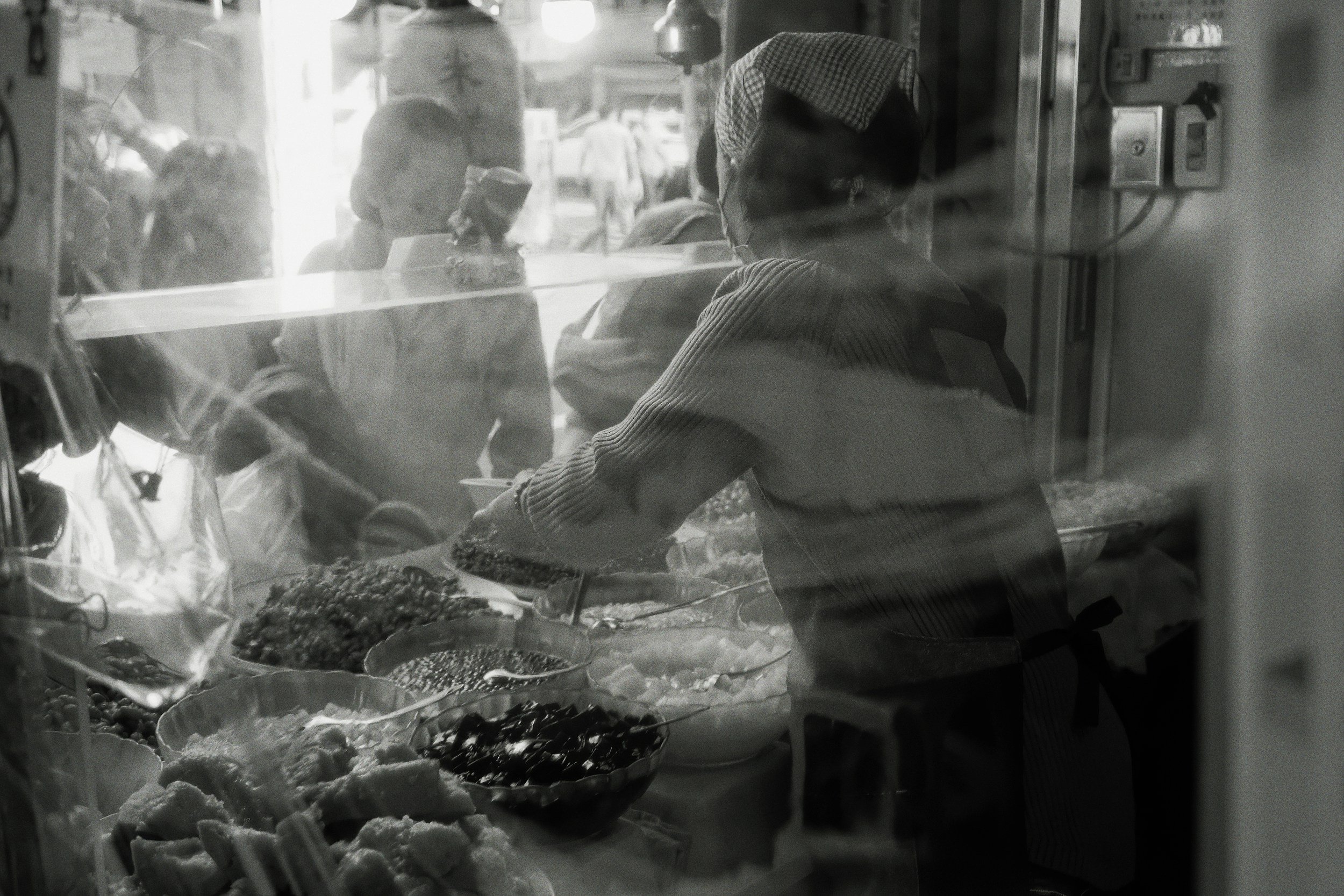A woman wearing a striped sweater and checkered headscarf preparing food behind a glass display counter at a street food stall, with bowls of ingredients inside. Outside, people are walking on the street.