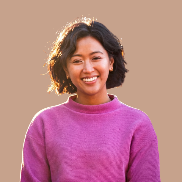 A young woman with short curly hair smiling, wearing a pink sweater, with a light brown background.