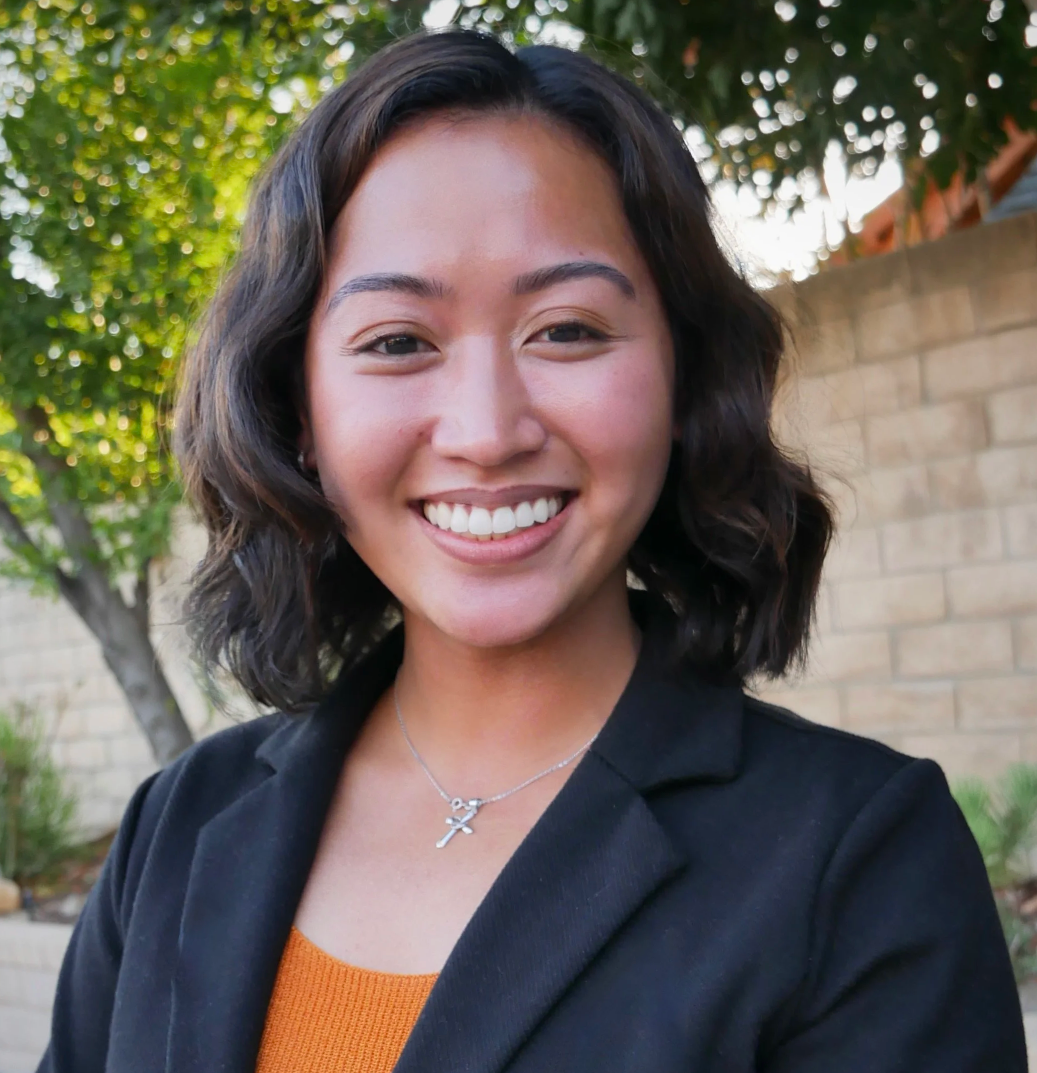 A smiling young woman with shoulder-length dark wavy hair outdoors, wearing a black blazer, an orange top, and a silver necklace with a cross pendant. There are trees and a brick wall in the background.