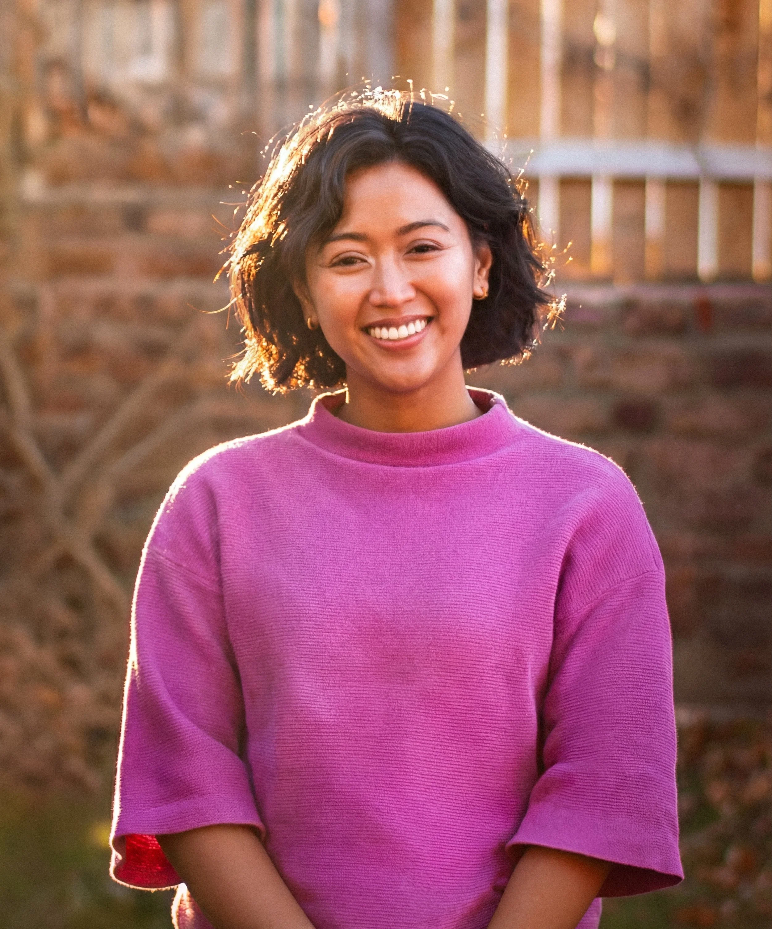Young woman with short curly hair smiling outdoors at sunset, wearing a pink sweater.