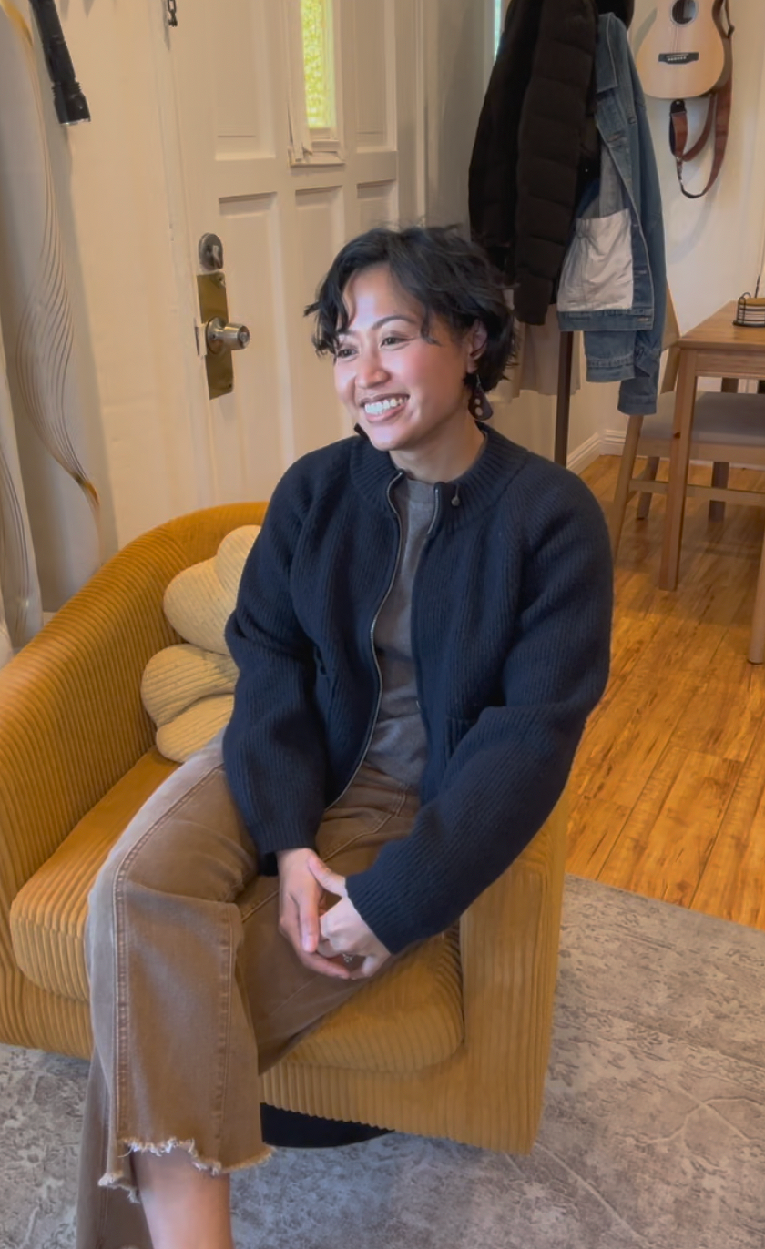 Helen Bass - A woman sitting on a yellow armchair in a cozy home interior, smiling, with a coat and guitar hanging on the wall behind her.