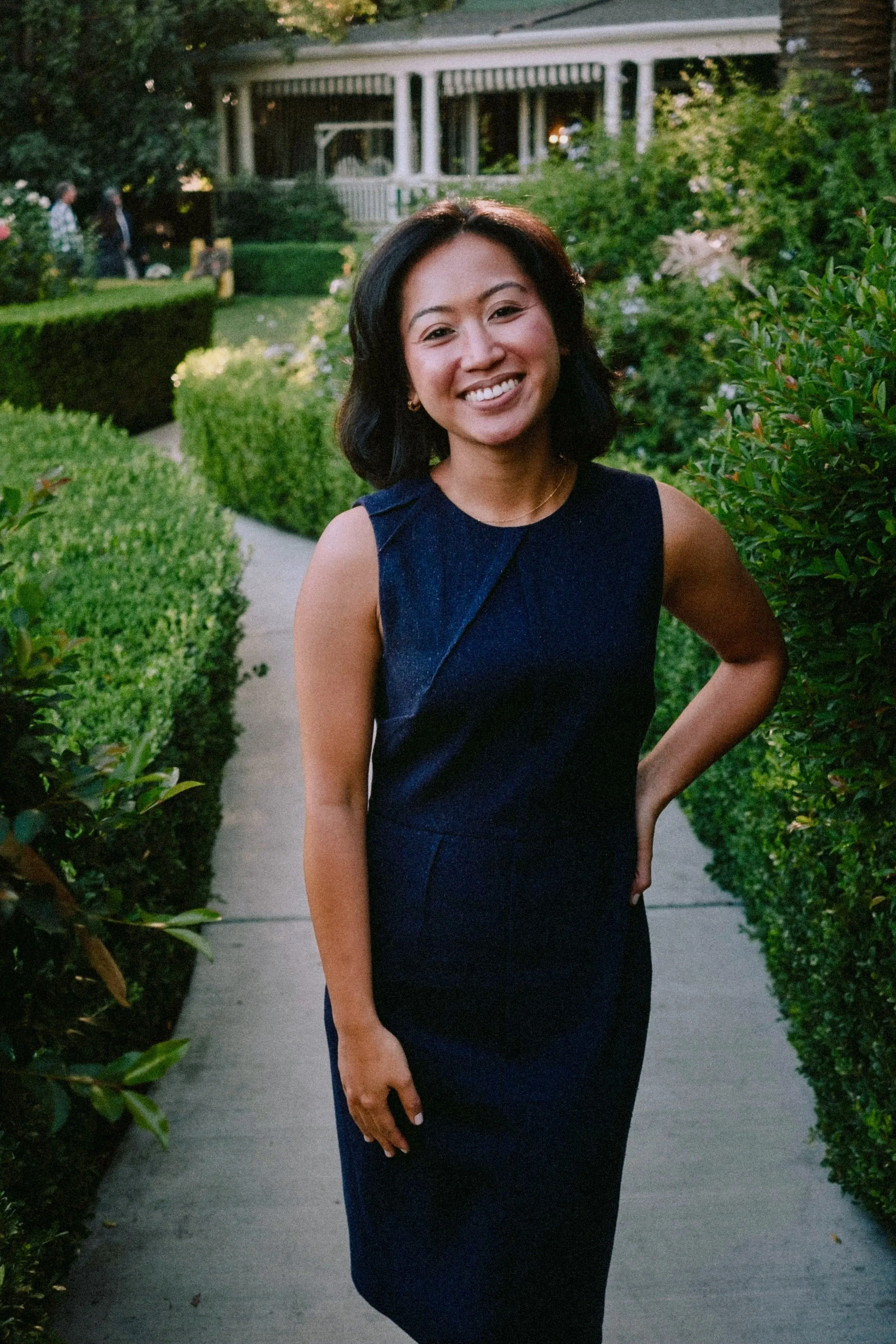 Helen Bass - A woman in a dark blue dress standing on a garden pathway, smiling, with greenery and a porch in the background.