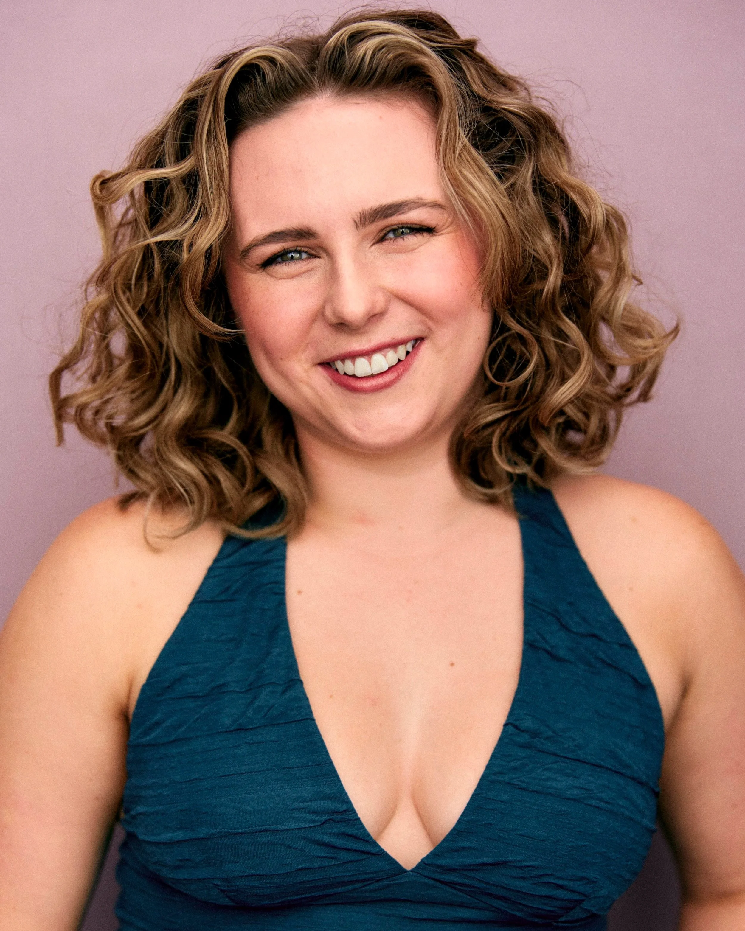 A woman with curly, shoulder-length hair smiling at the camera, wearing a dark blue sleeveless top with a textured fabric, against a light purple background.