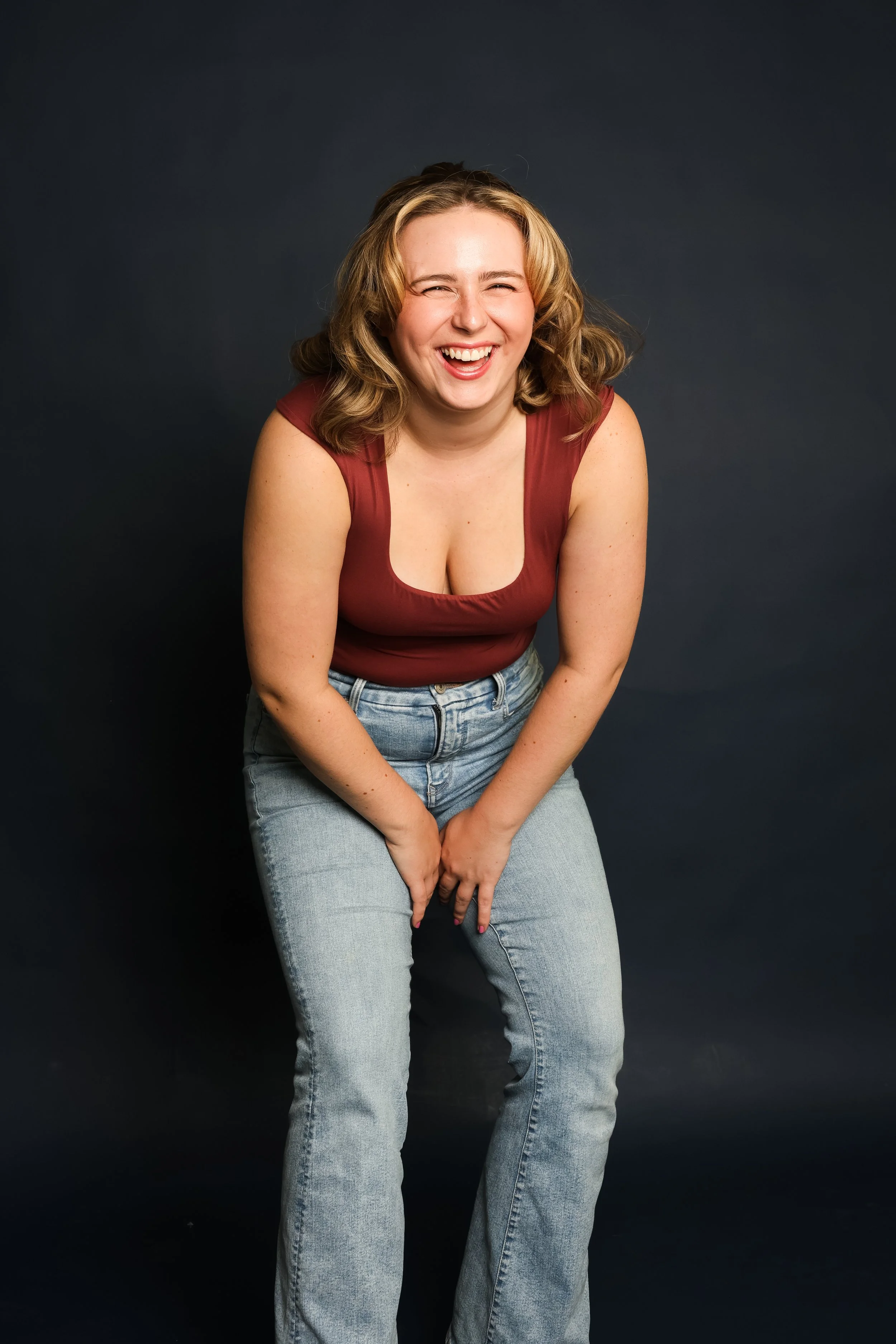 A woman with curly blonde hair wearing a sleeveless maroon top and light blue jeans, laughing and leaning forward against a dark background.