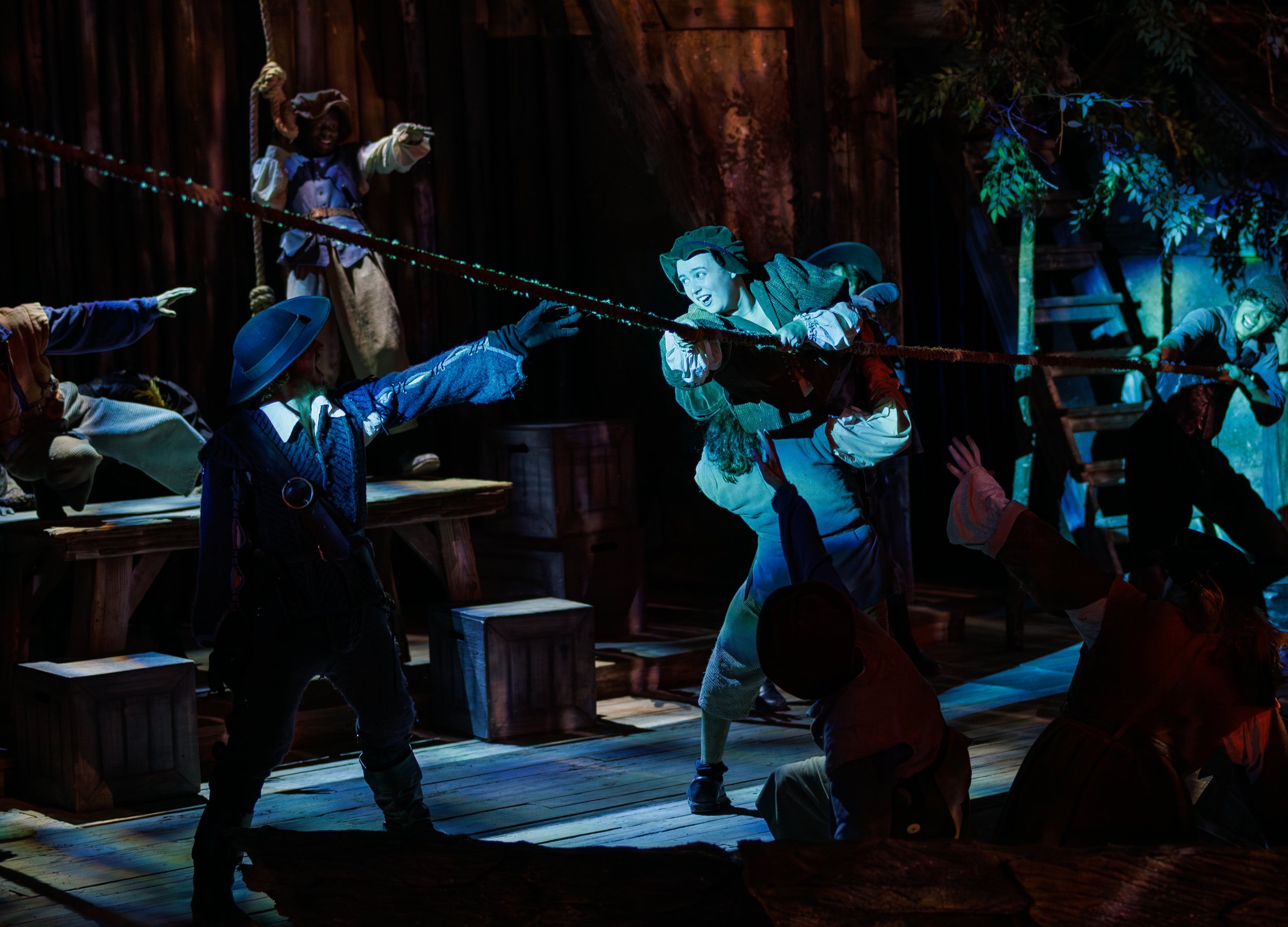 The image shows a theatrical performance of a scene from 'Peter Pan' with actors dressed as characters from the story, including Peter Pan. They are engaged in a mock sword fight on stage with dim lighting and a forest-themed background.