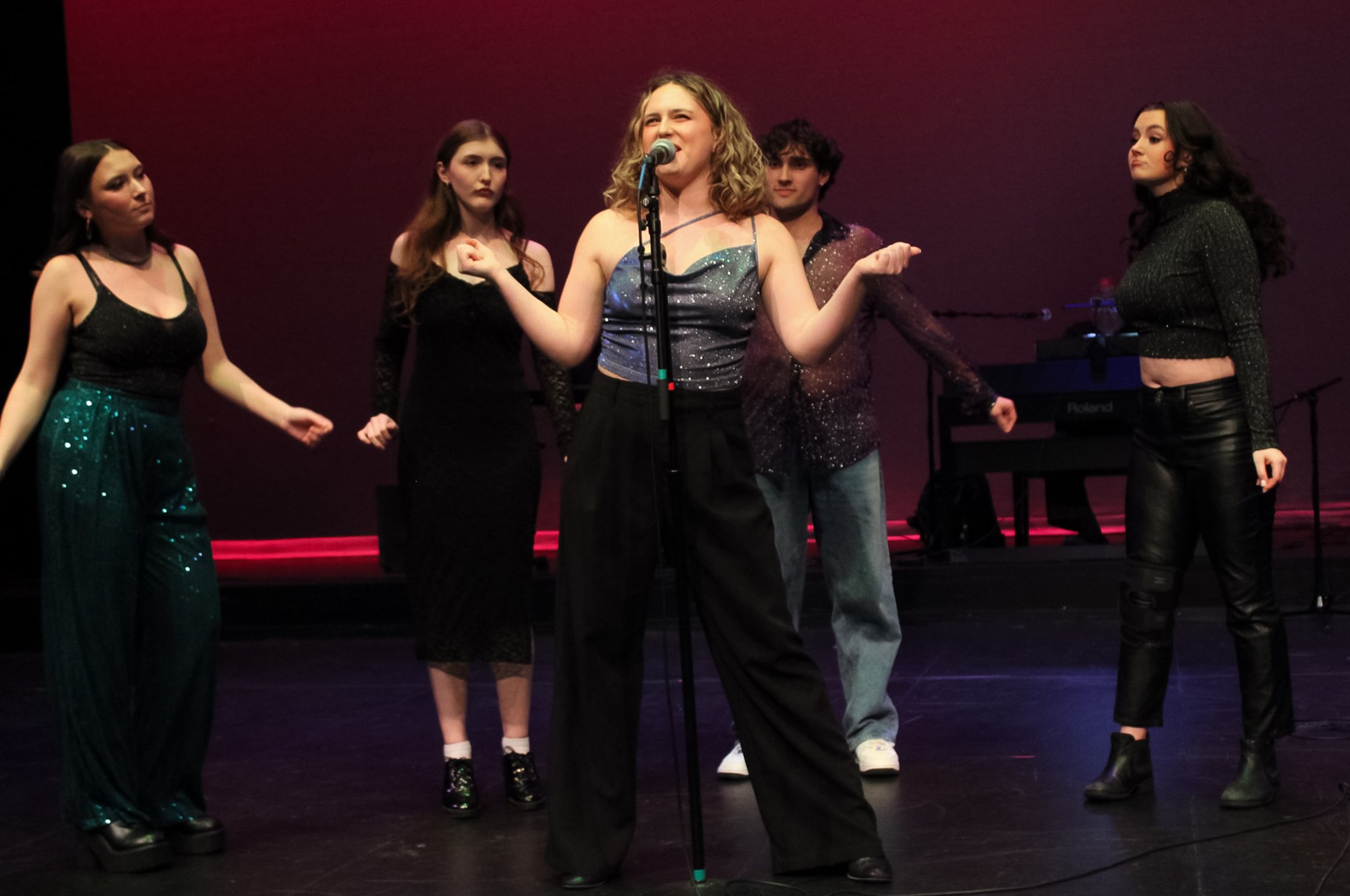 Group of five young women standing on stage, performing, with a pinkish-red background and a keyboard in the background.
