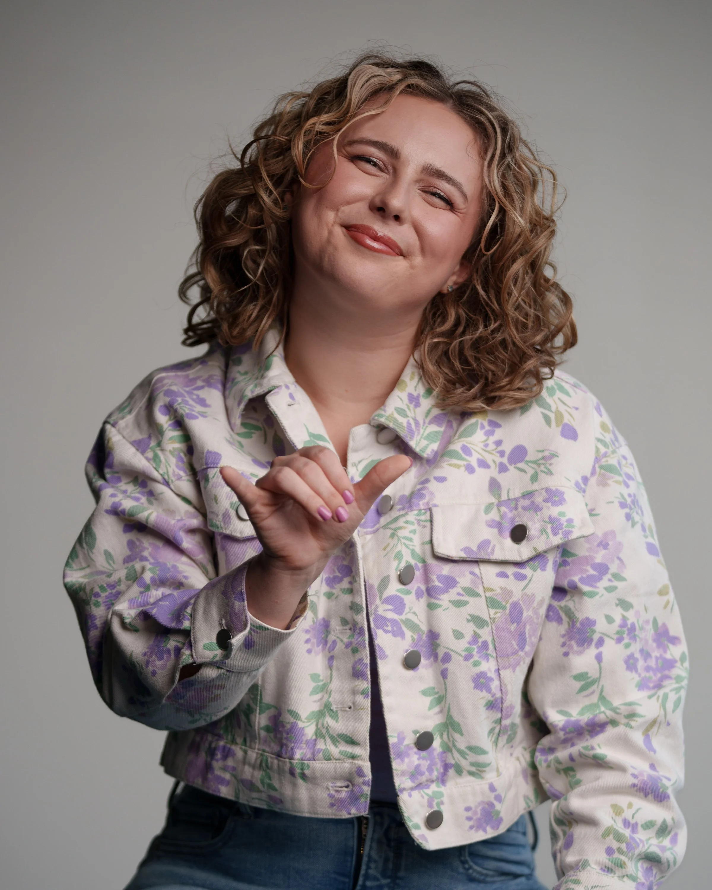 A young woman with curly blonde hair smiling and making a shaka hand gesture, wearing a floral-patterned denim jacket.