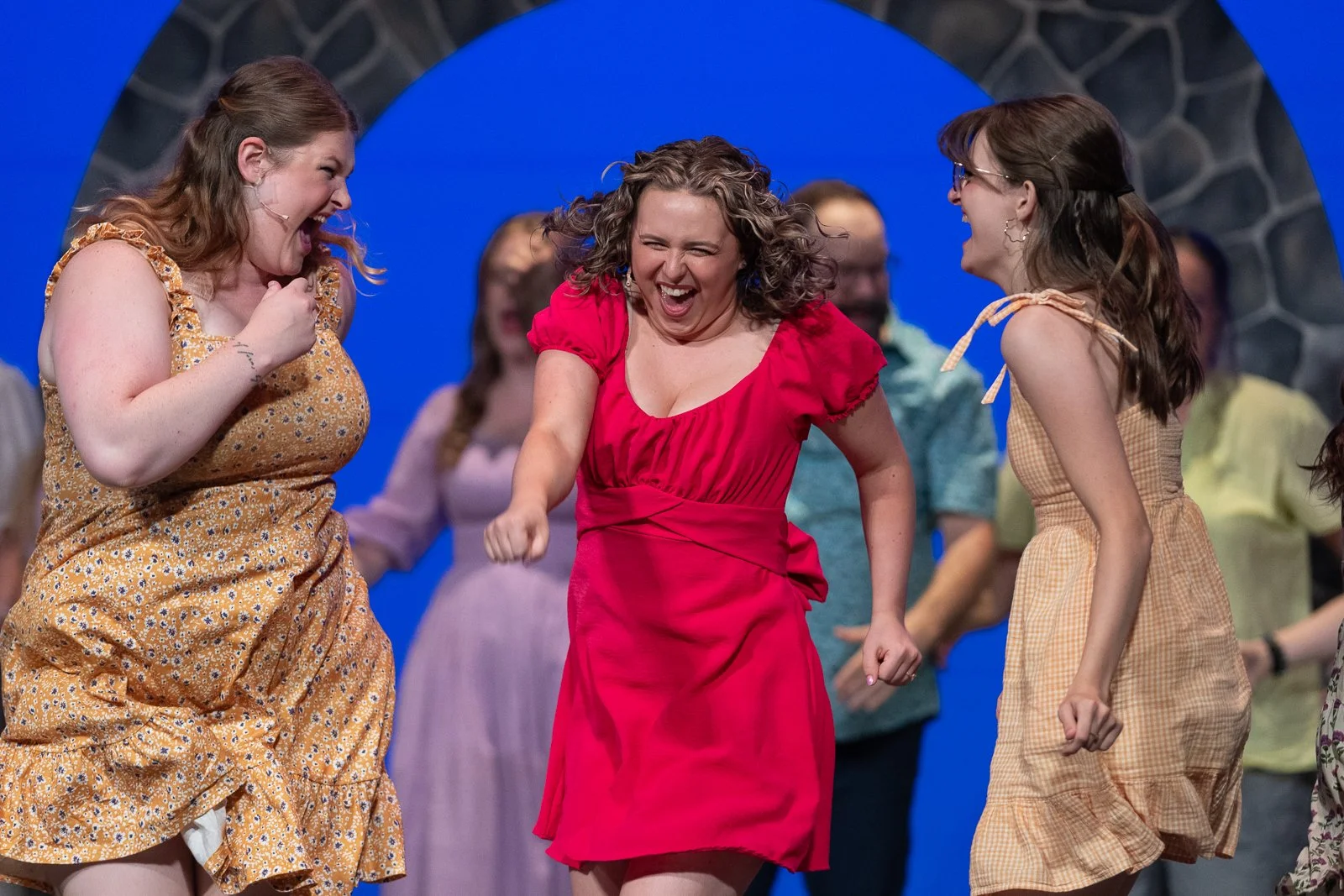 Three women laughing and celebrating on stage with a blue background, dressed in colorful dresses.