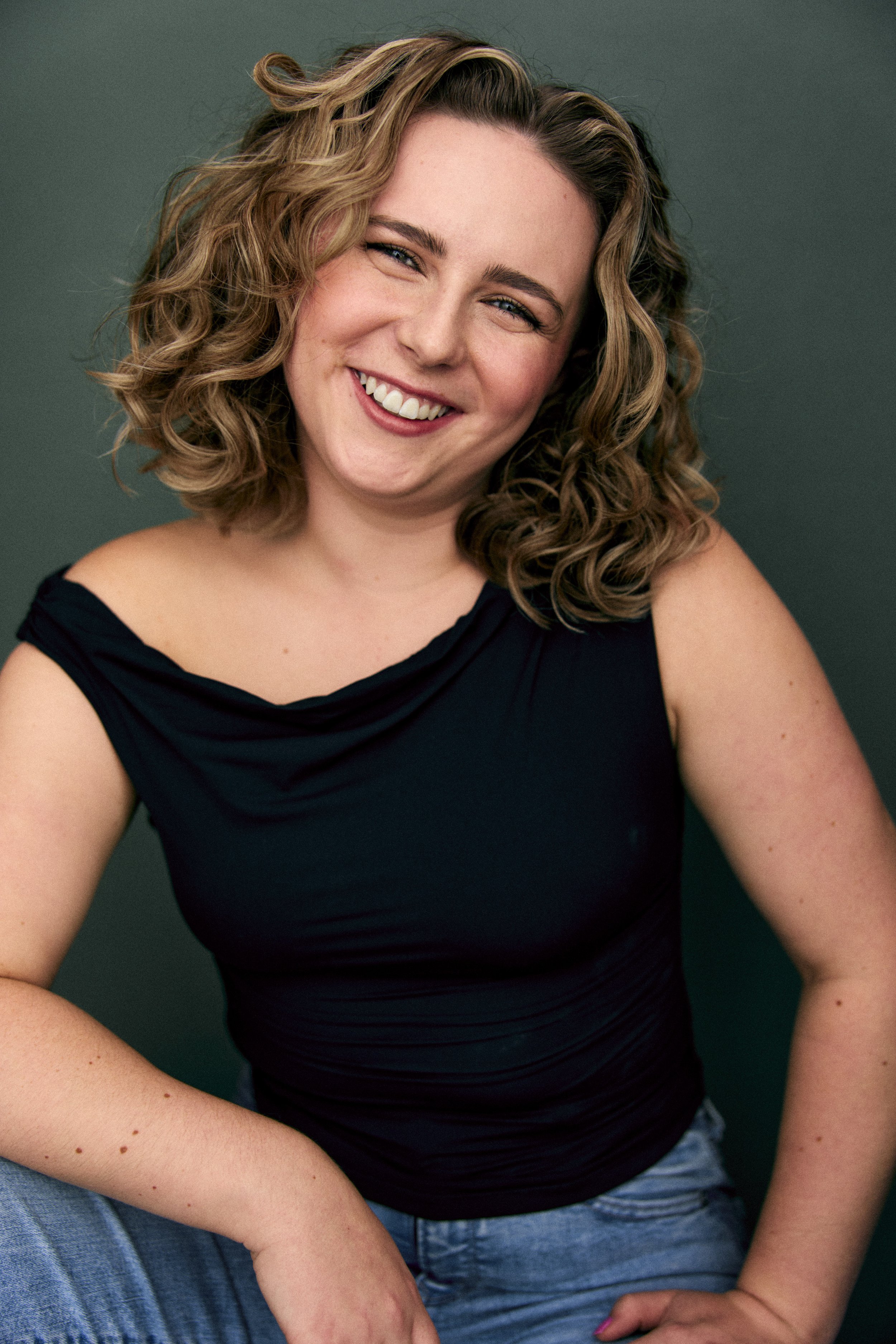 A smiling woman with curly blonde hair, wearing a black sleeveless top and blue jeans, sitting against a dark green background.