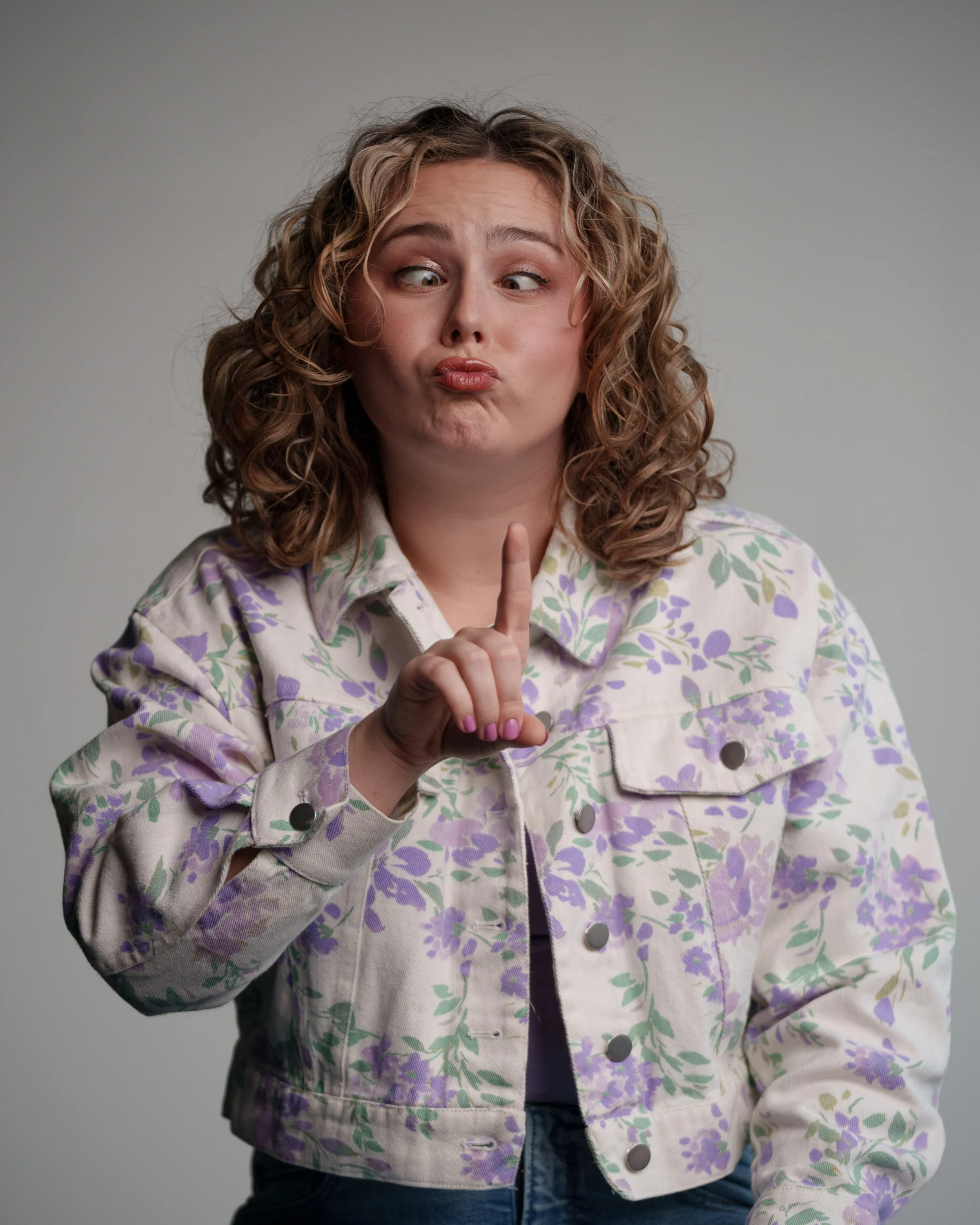 Young woman with curly hair making a duck face and holding her index finger up in a gesture of emphasis, wearing a floral patterned denim jacket.