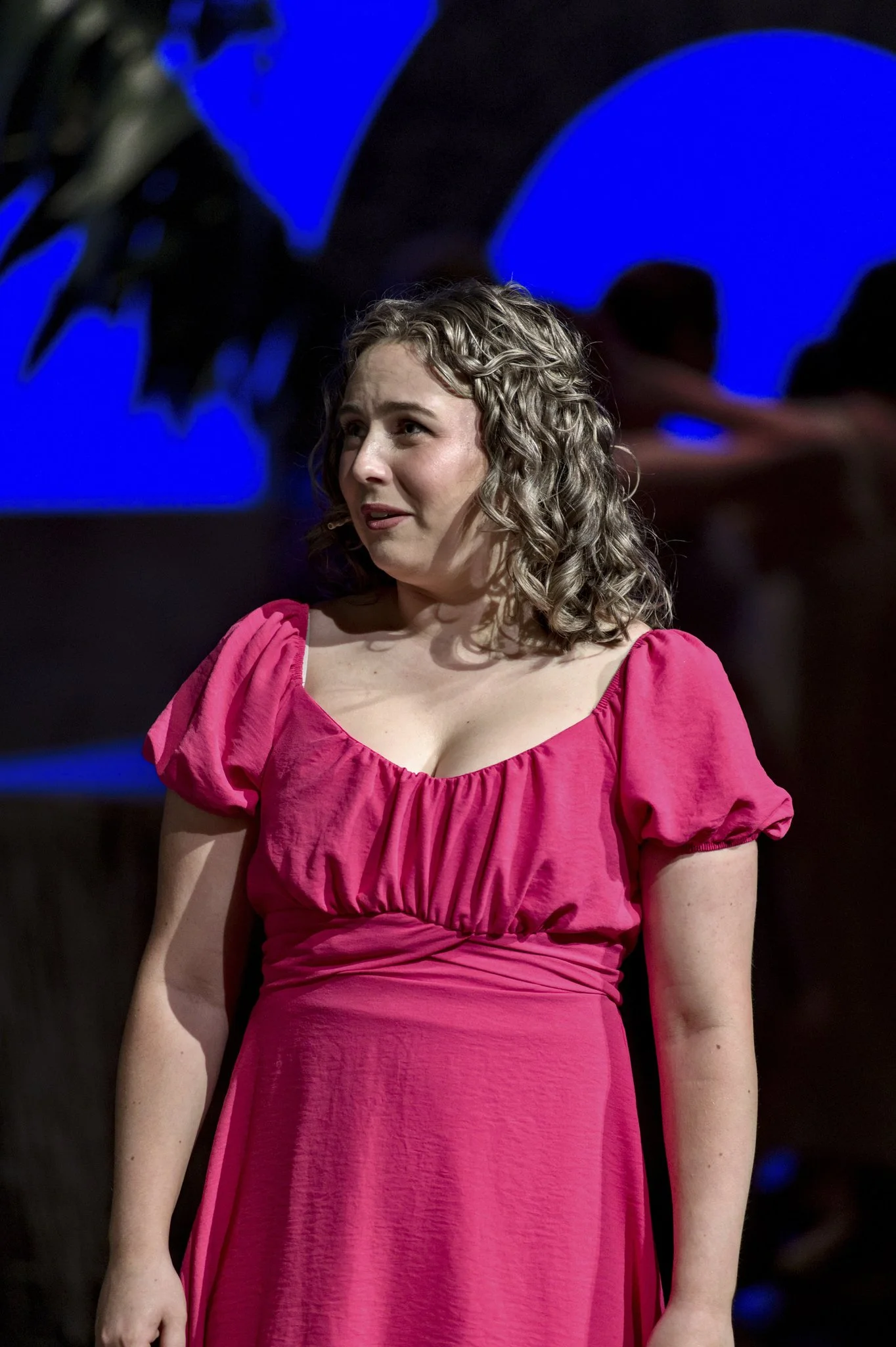 A young woman with curly hair wearing a pink dress standing on stage with a blue background.