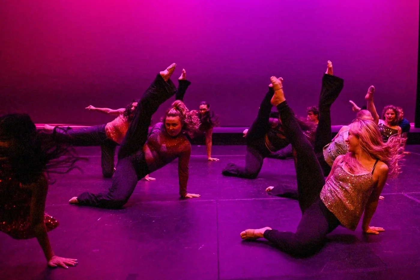 Group of women performing a dance routine on stage with purple lighting, some with legs raised, all in dance poses.
