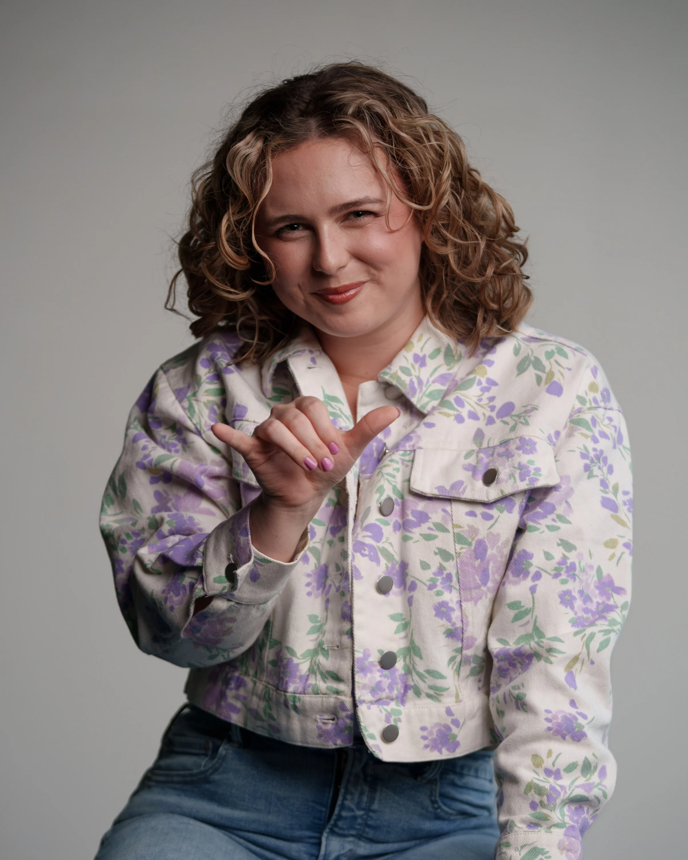 A woman with curly, shoulder-length hair, wearing a floral patterned jacket, making a shaka sign with her right hand and smiling playfully.