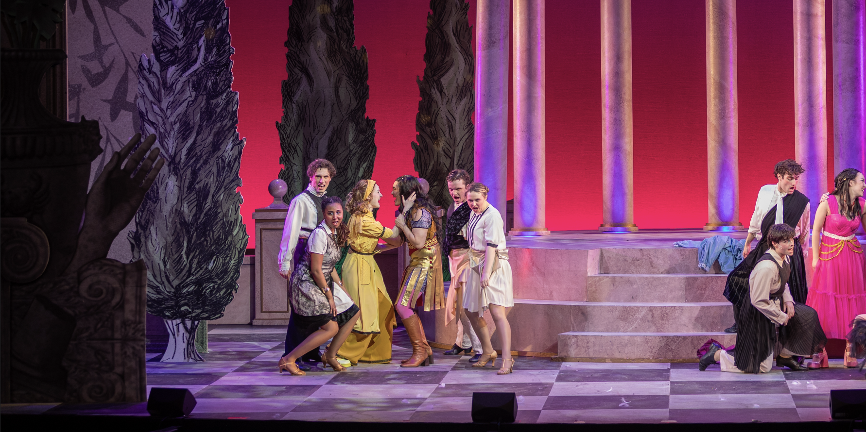 Theatrical stage with actors in colorful costumes portraying a lively scene involving multiple women and men, some engaged in conversation, others kneeling or standing, amid set pieces resembling trees and columns with a pink and purple backdrop.