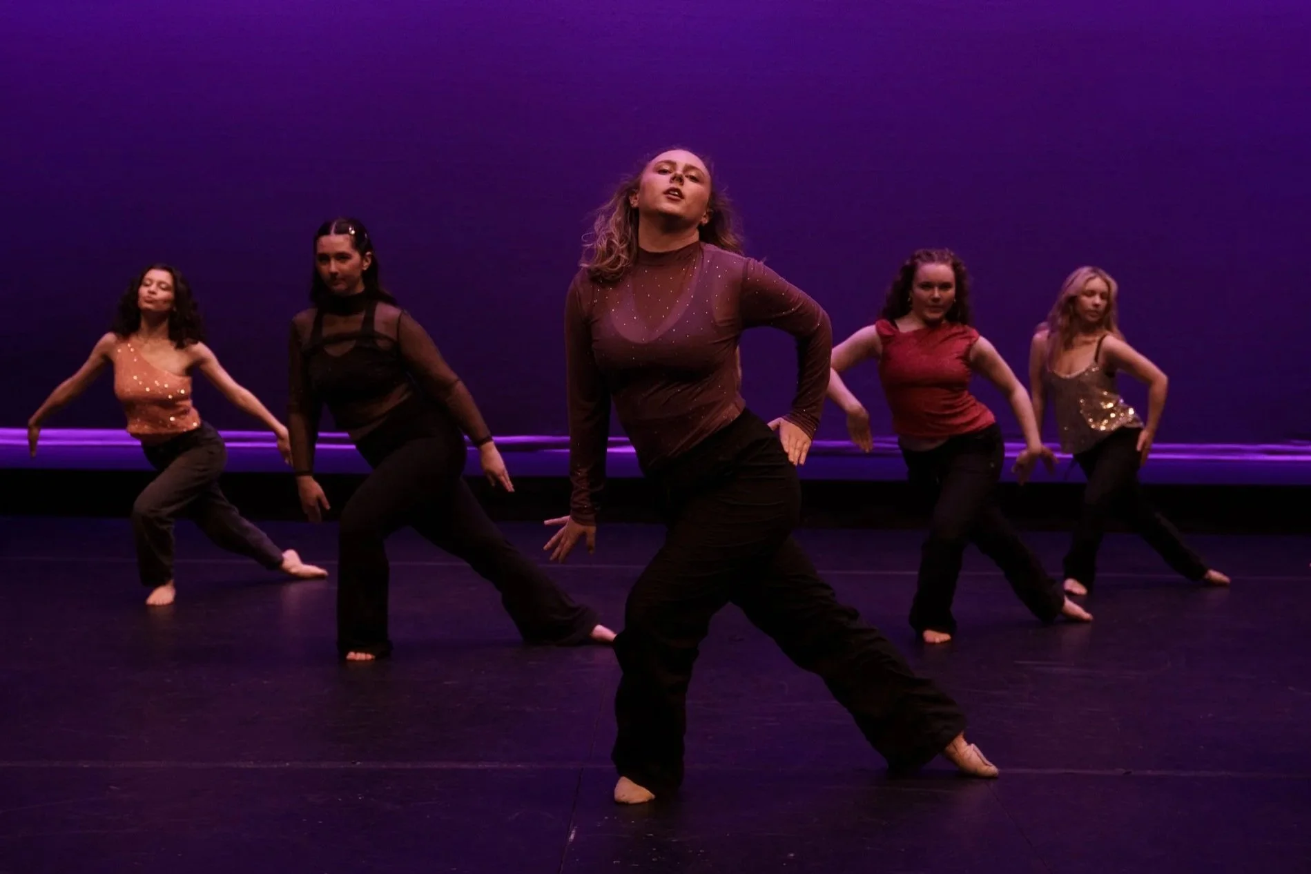 Five women dance on stage with purple lighting in the background.