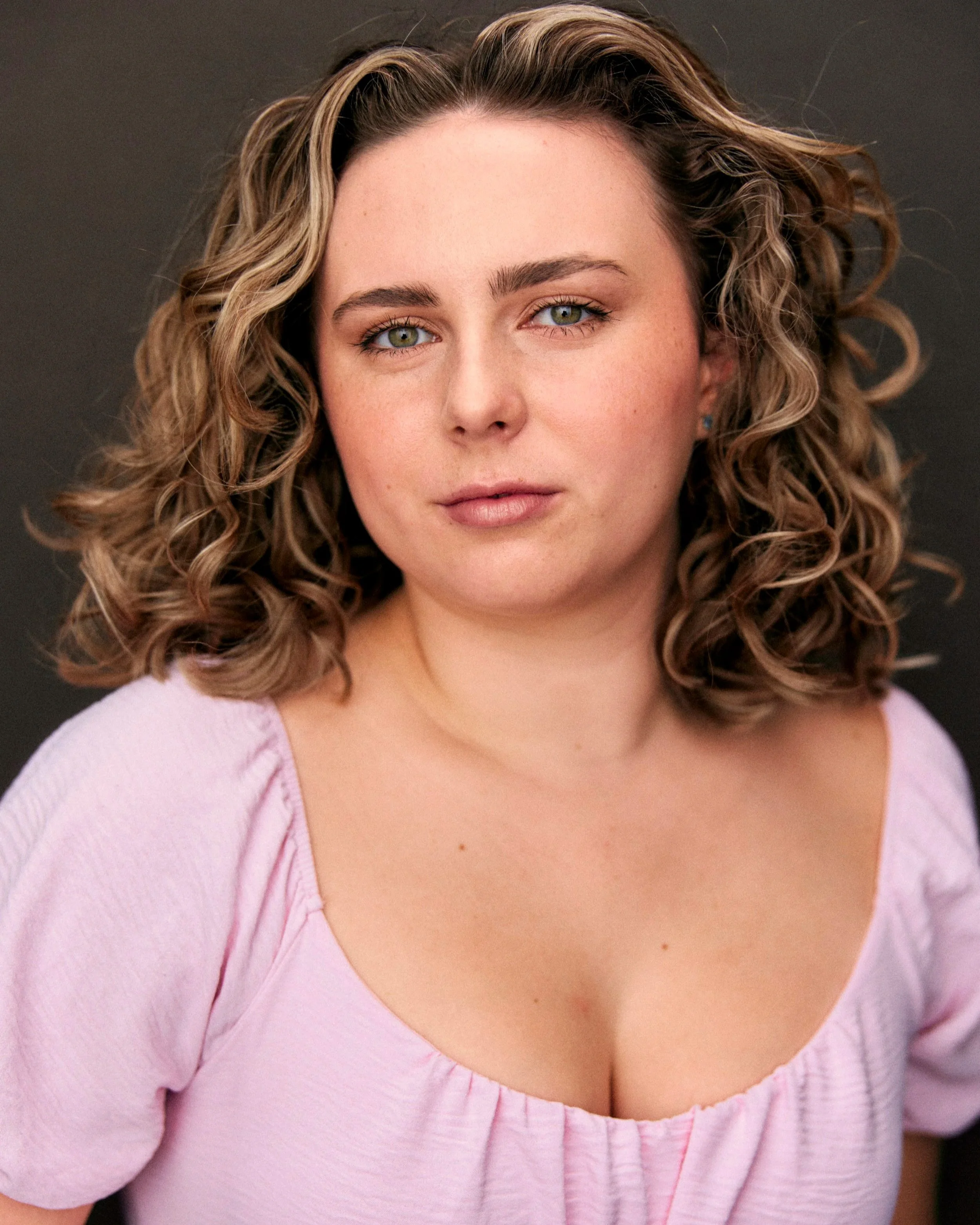 A woman with shoulder-length curly brown hair, wearing a pink top, looking directly at the camera with a neutral expression against a dark gray background.