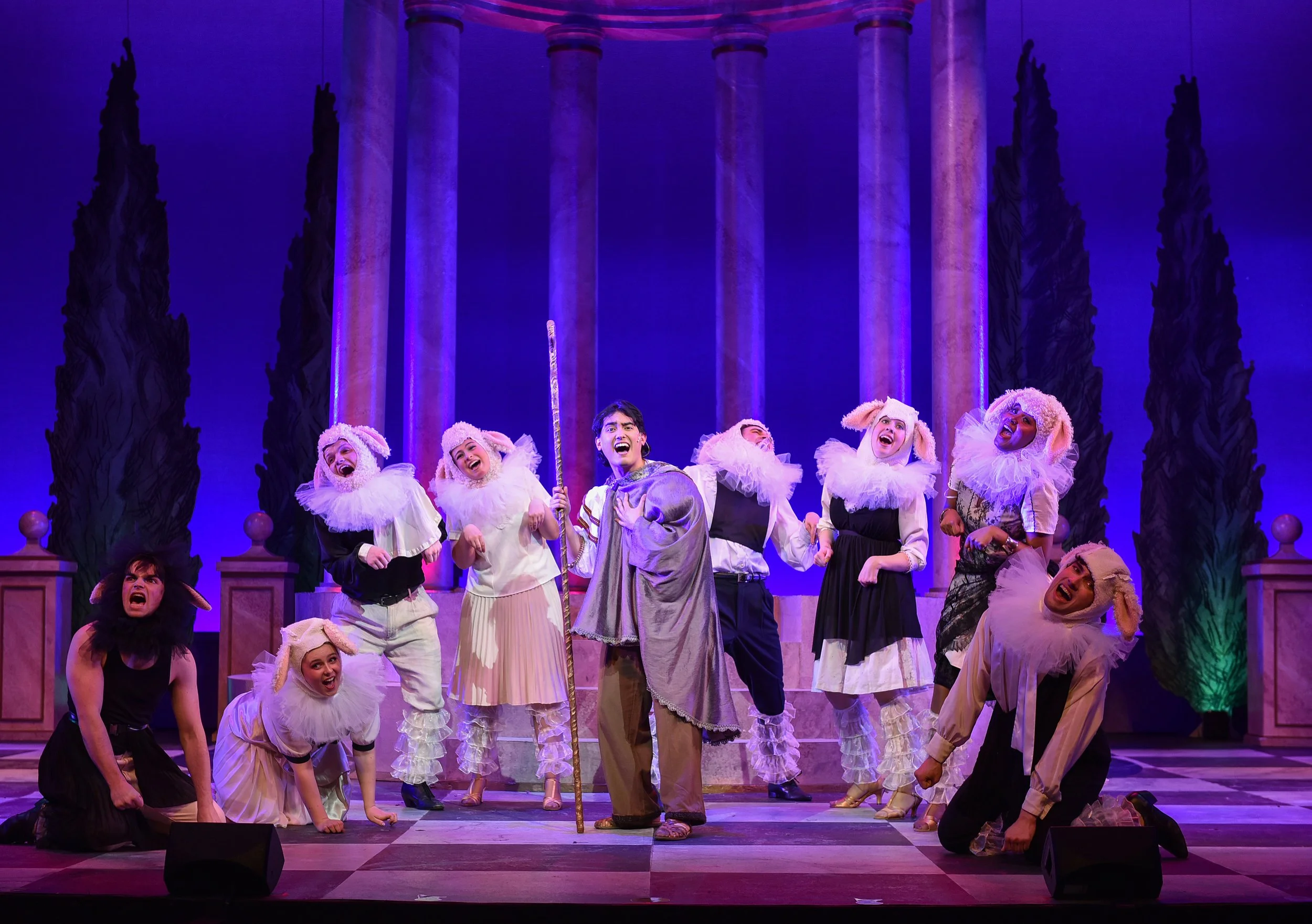 A theatrical stage performance featuring actors dressed as sheep with white fluffy costumes and a person dressed as a lion with a black costume and mane. The scene includes a central character holding a staff, possibly portraying a leader or storyteller. The background has tall columns and dark trees, with vibrant lighting creating a lively atmosphere.