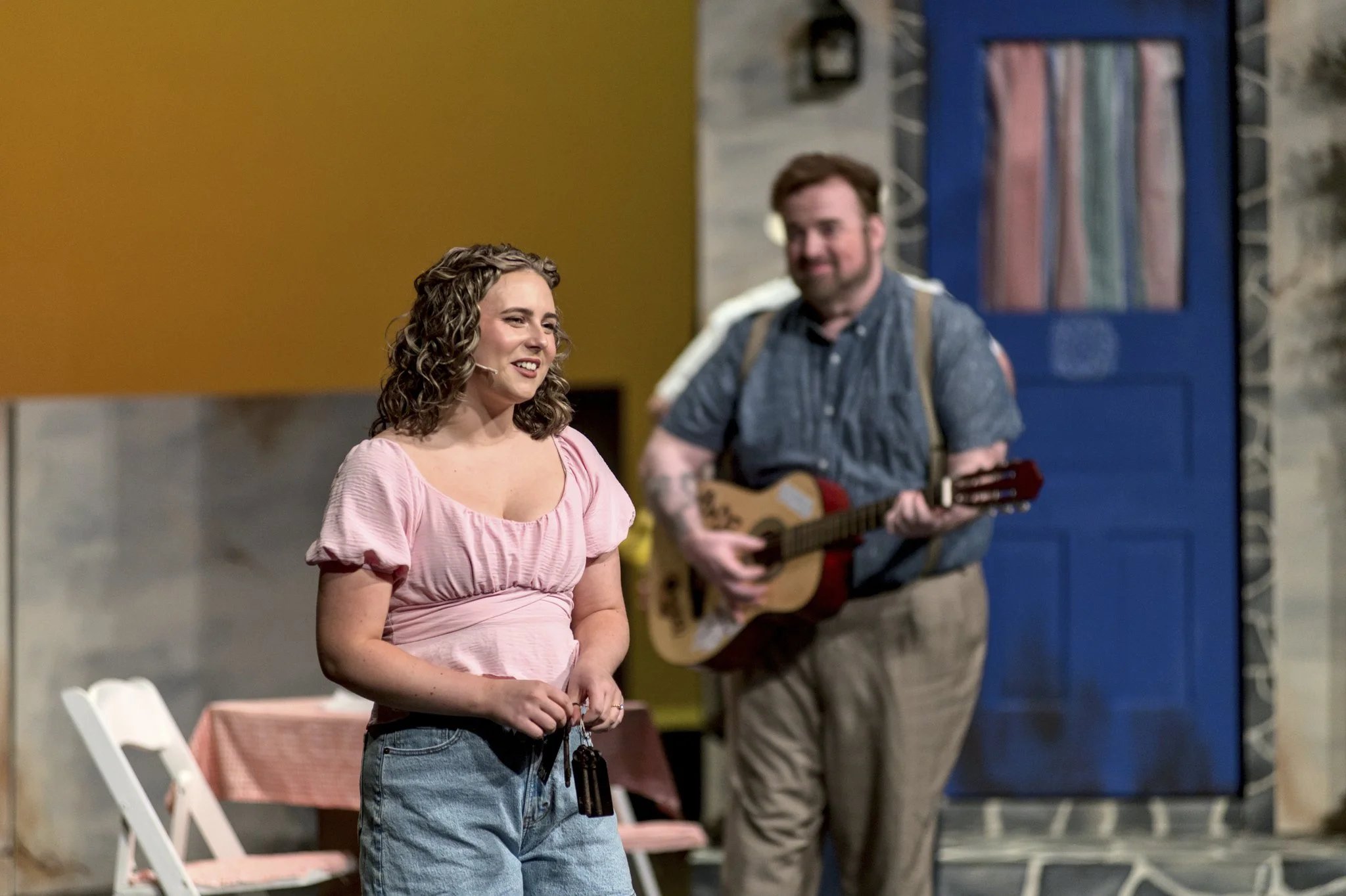 A woman in a pink top and jeans holding keys stands in front of a man playing a guitar on a stage set, with a blue door and colorful curtains in the background.