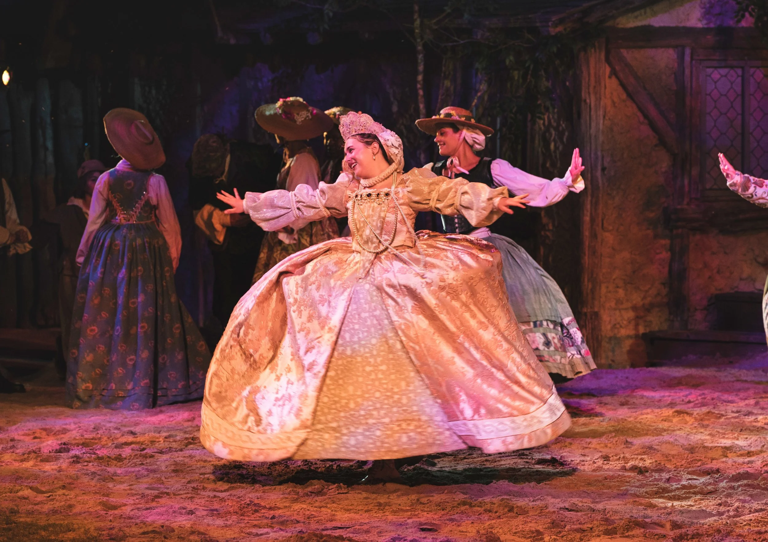 A woman in a fancy, old-fashioned gown dances with her arms outstretched, surrounded by other women in similar period costumes on a stage with warm lighting and rustic background.