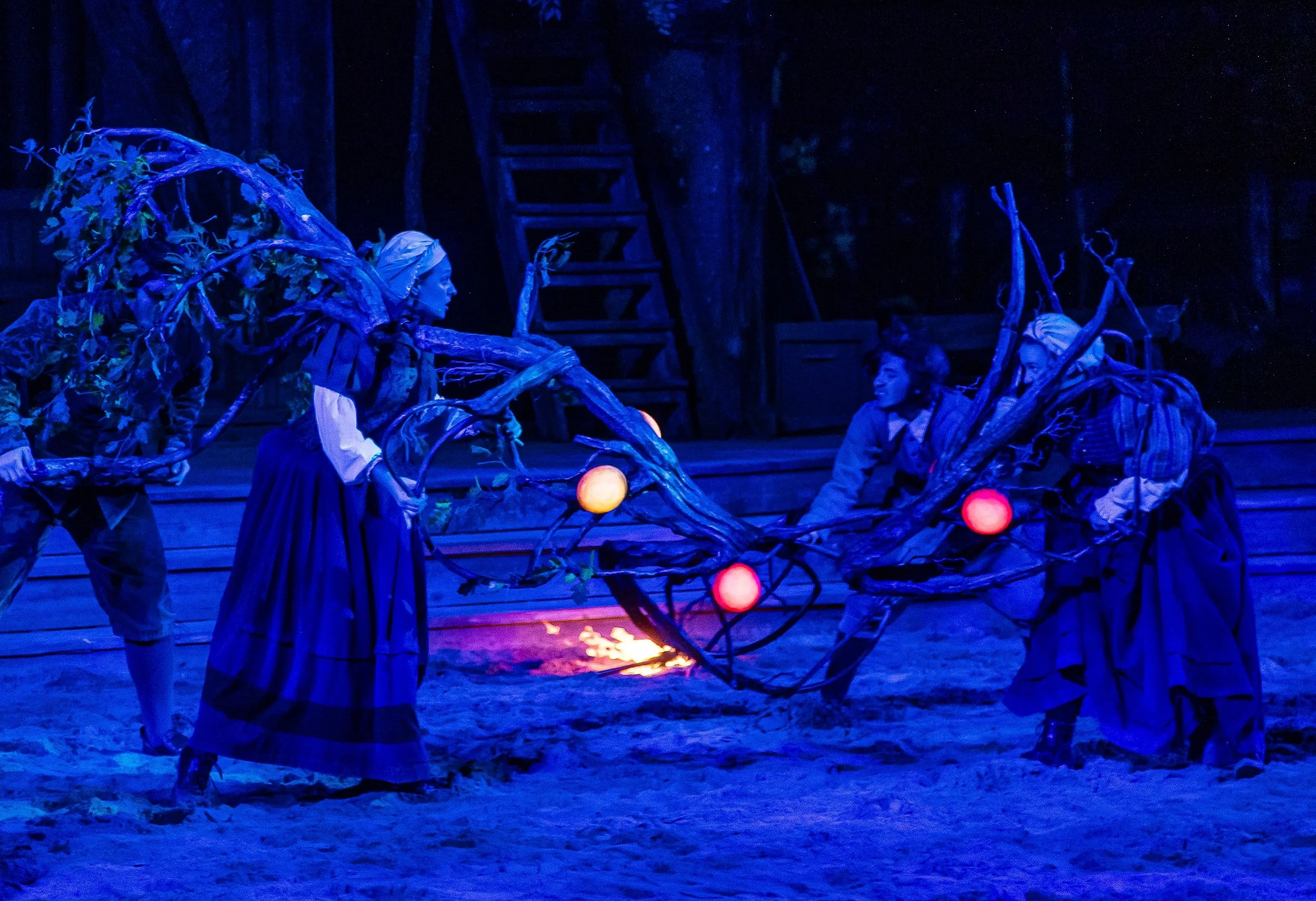The image shows a theatrical performance with actors dressed as trees or plant characters, with glowing orbs connected by branches. A woman in period costume is reaching toward a person on the ground, in a dimly lit setting with blue and purple lighting, on a stage with wooden elements in the background.