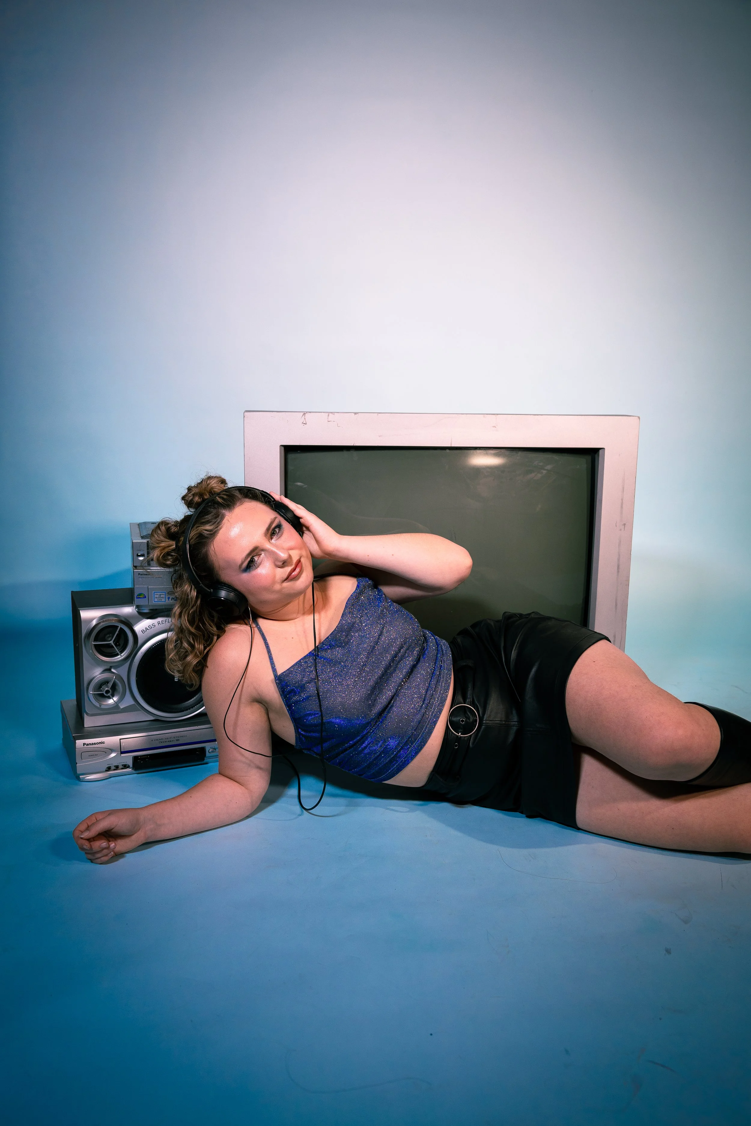 A young woman in a sparkly blue crop top and black shorts lying on a blue studio floor, wearing headphones and resting against a large TV, with a stereo system nearby.