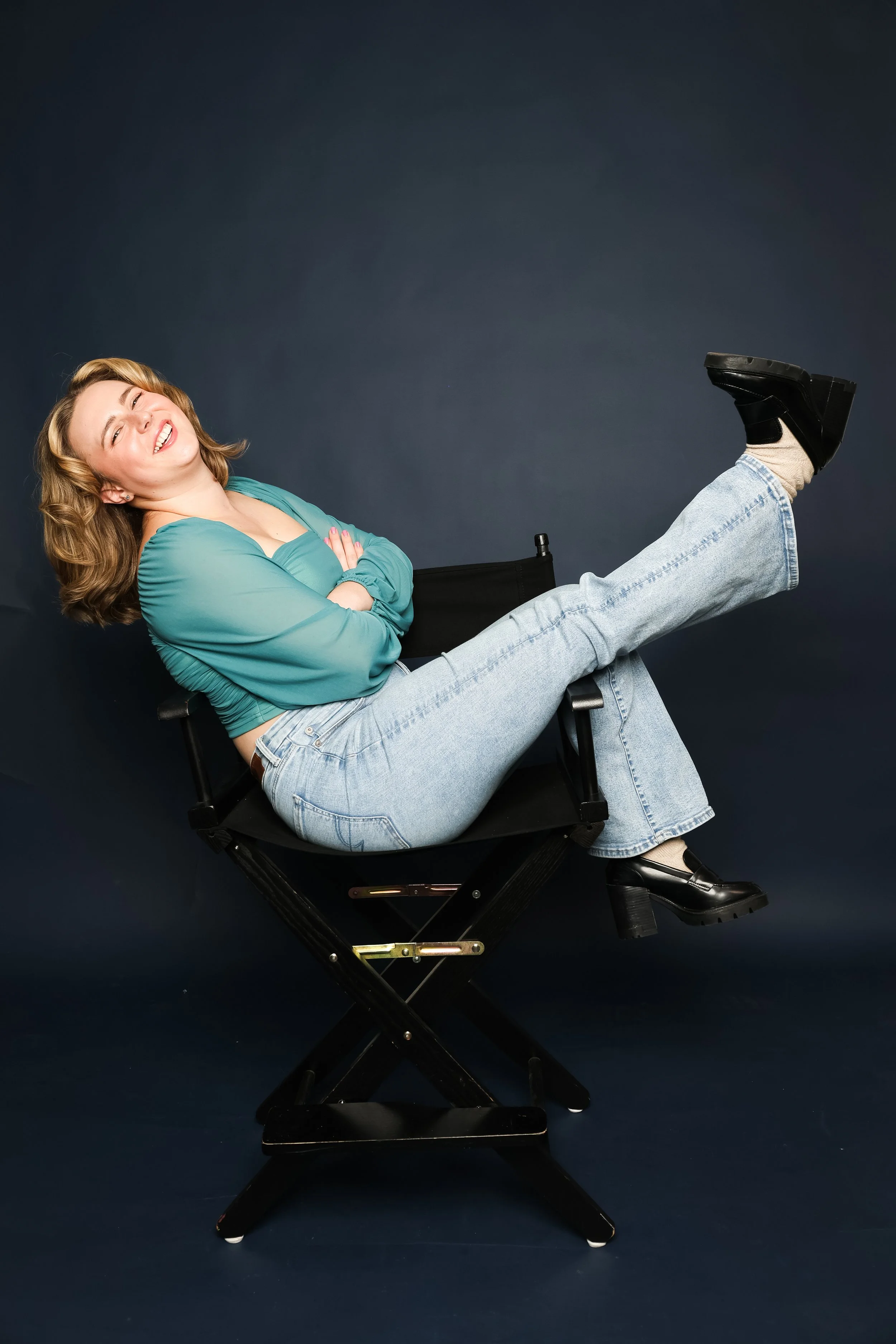 Woman sitting in a director's chair, smiling with arms crossed, wearing a teal top and light blue jeans, with one leg raised and black shoes, against a dark background.