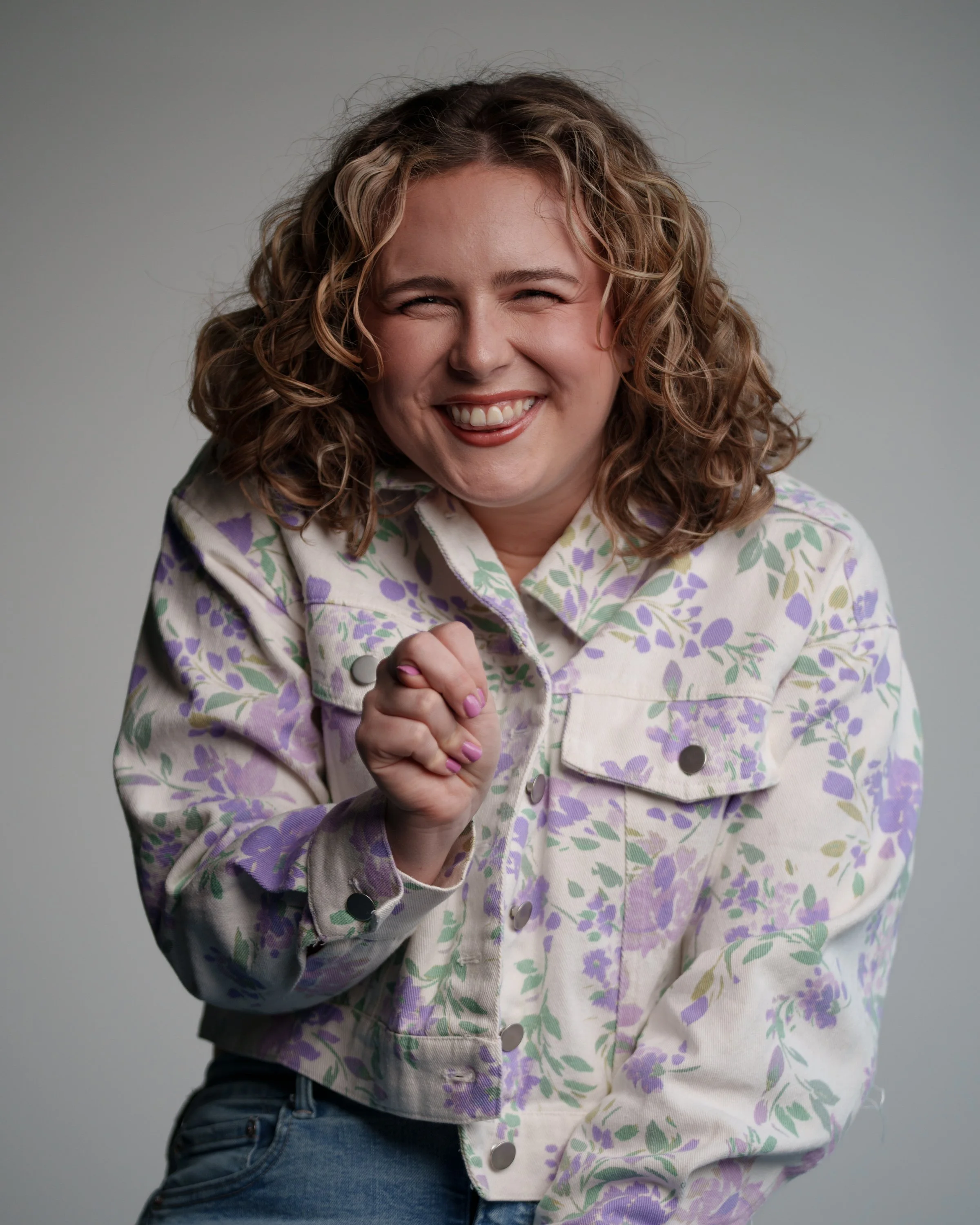 A woman with curly hair smiling and making a fist gesture, wearing a floral jacket.