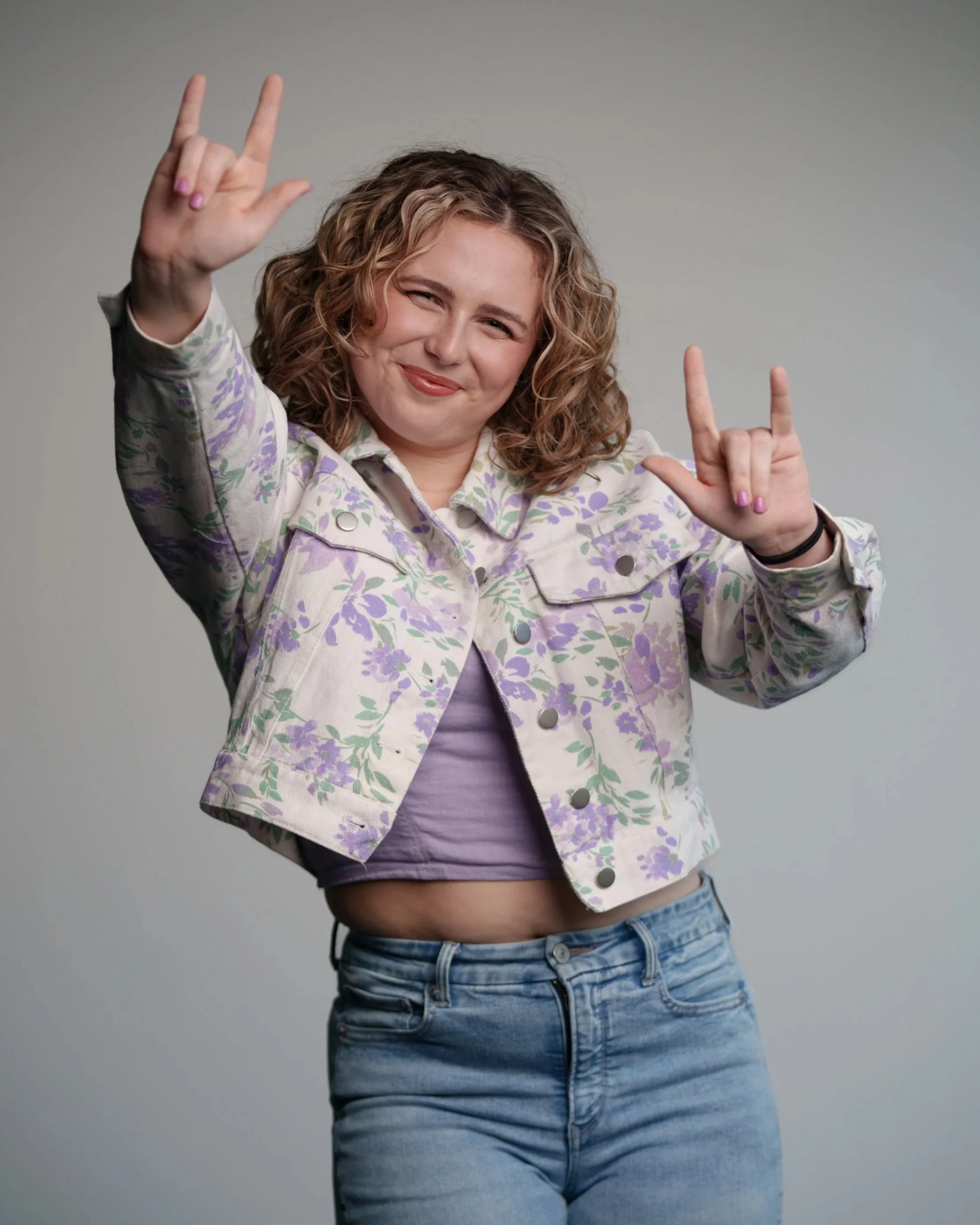 A young woman with curly blonde hair making rock and roll hand gestures, smiling, wearing a floral jacket, purple shirt, and jeans against a plain gray background.