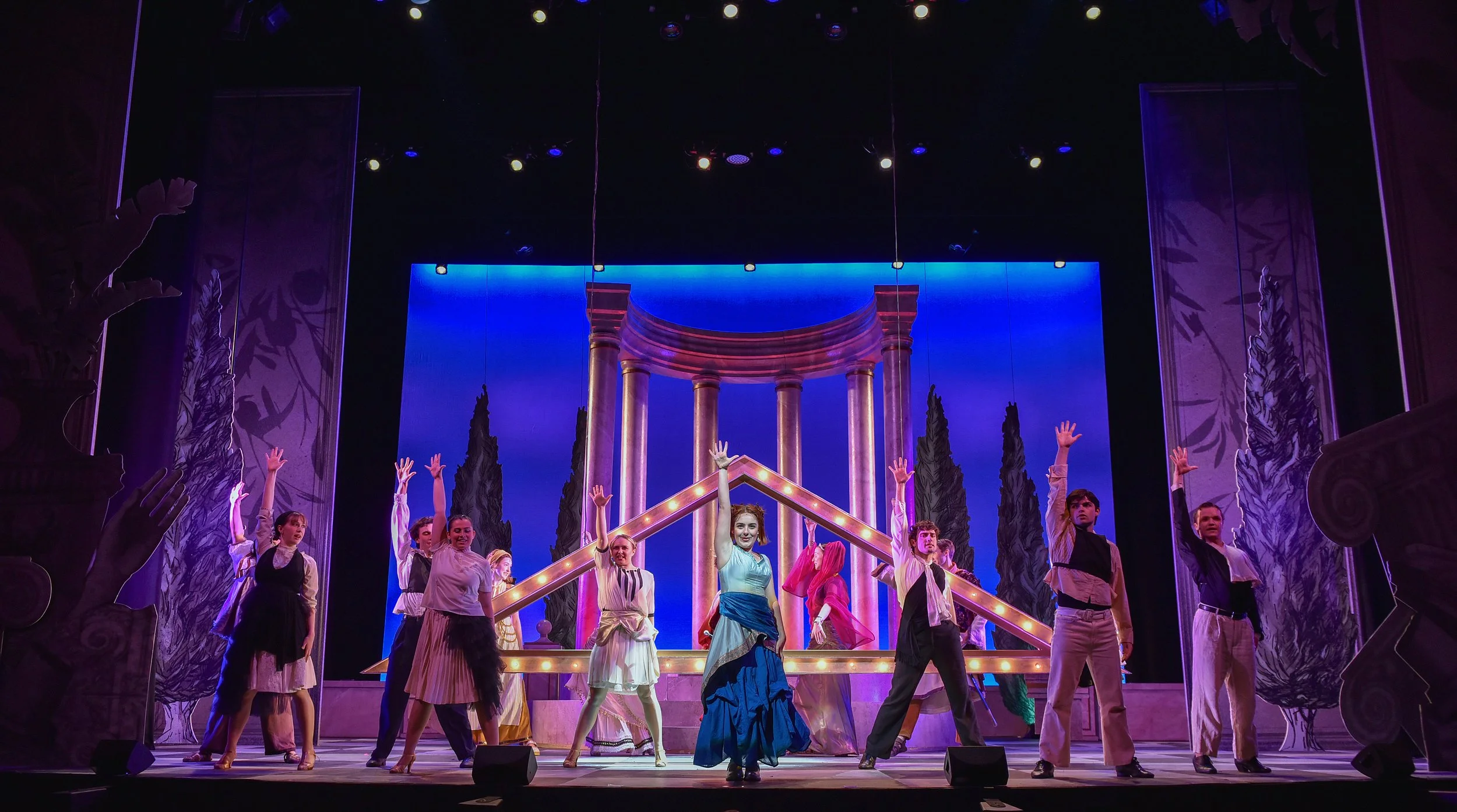 A theater stage with wooden columns and a blue backdrop featuring tall trees and a structure resembling a classical temple. A group of performers in colorful costumes are dancing with their arms raised.