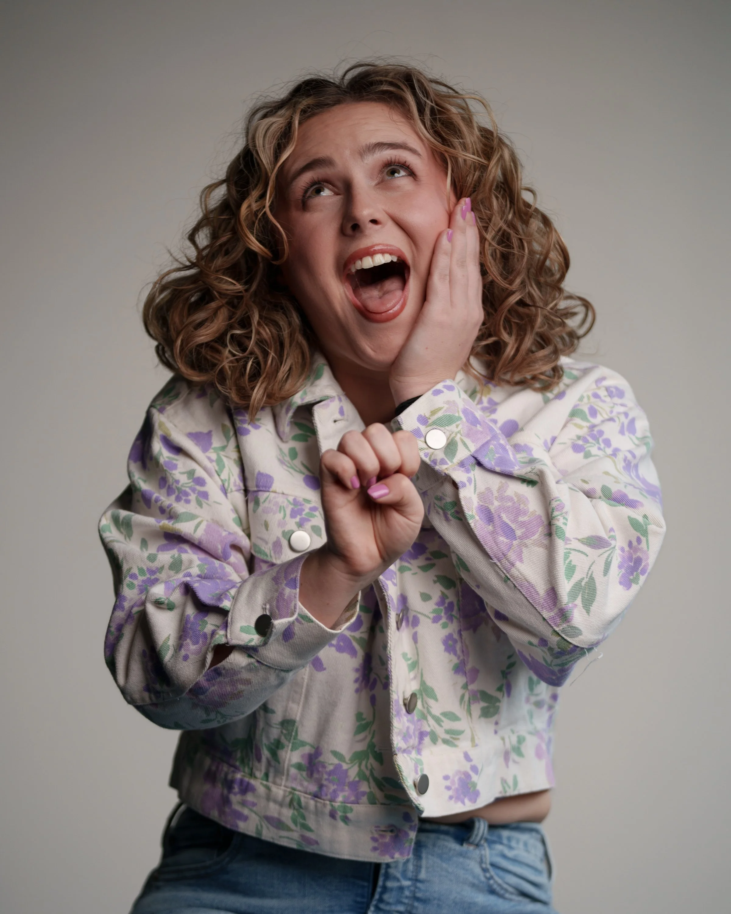 A woman with curly hair shows surprise and excitement, wearing a floral patterned jacket and blue jeans.