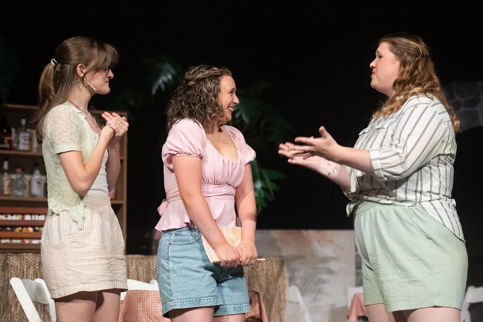 Three women having a conversation indoors, one is gesturing with hands, the other two are smiling, in a room with greenery and shelves.