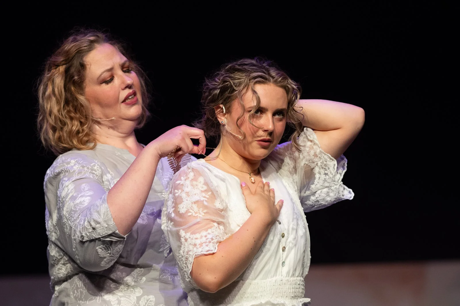 Two women on stage, one is helping the other with her hair, both wearing white embroidered dresses. The woman on the right has her hand on her chest and looks surprised or emotional.
