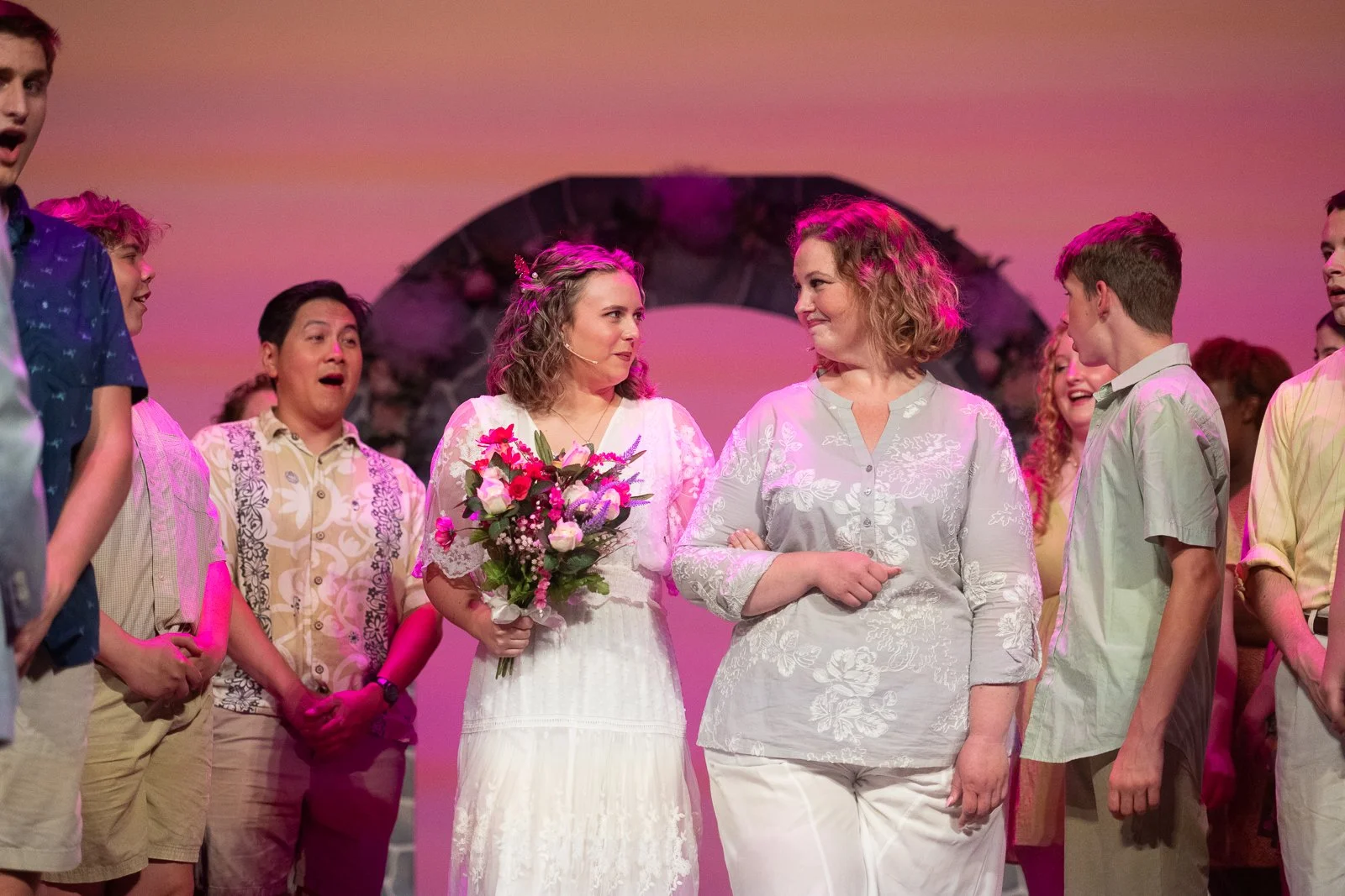 Stage scene with woman in white dress holding a bouquet of flowers and woman in light blouse, surrounded by appreciative young people, colorful background
