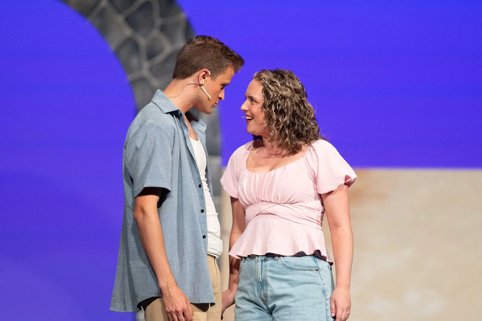 Two actors stand close together on a stage with a purple and light beige background, looking into each other's eyes and smiling.