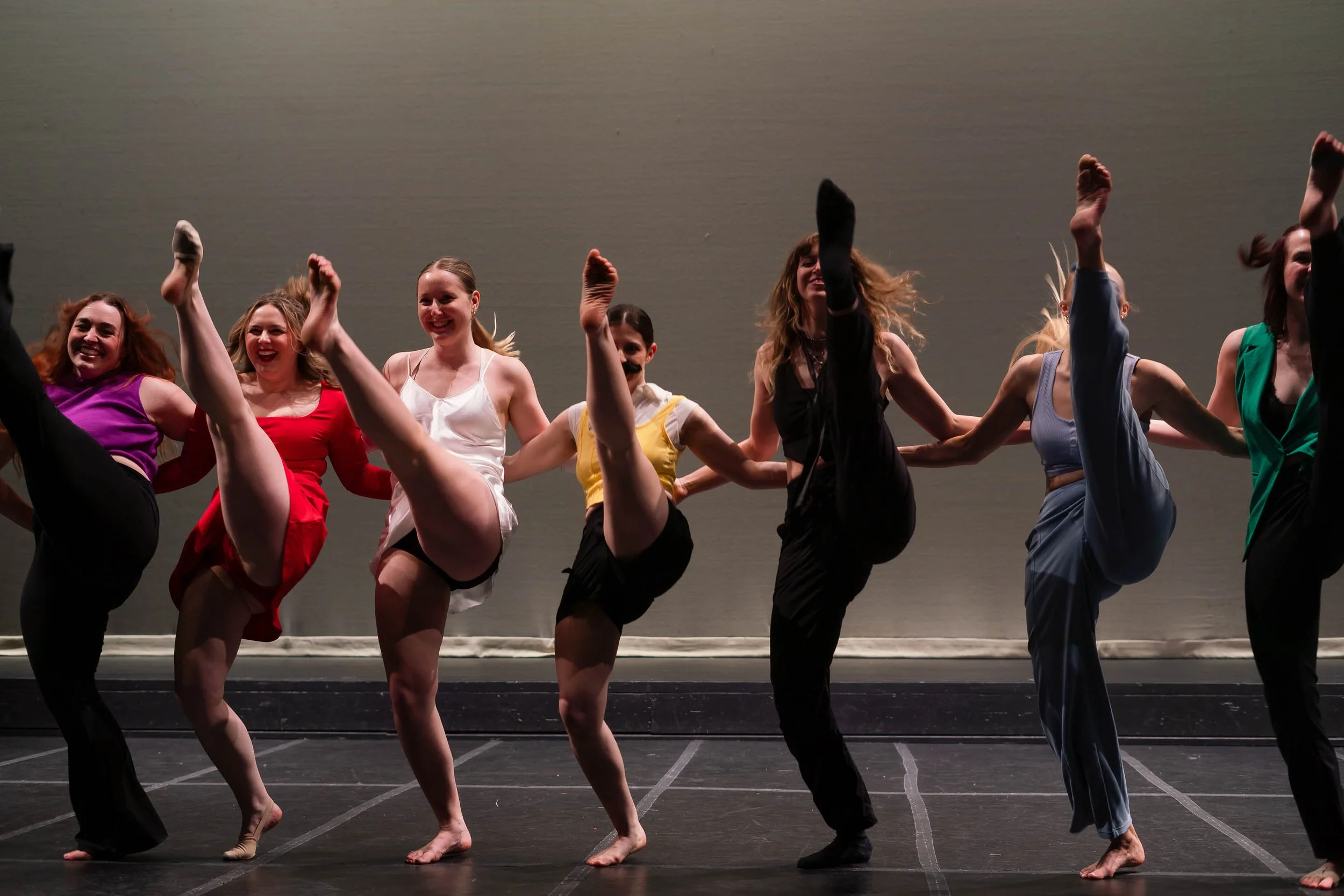 A group of women dancing on stage, holding hands, with one leg lifted high in the air.