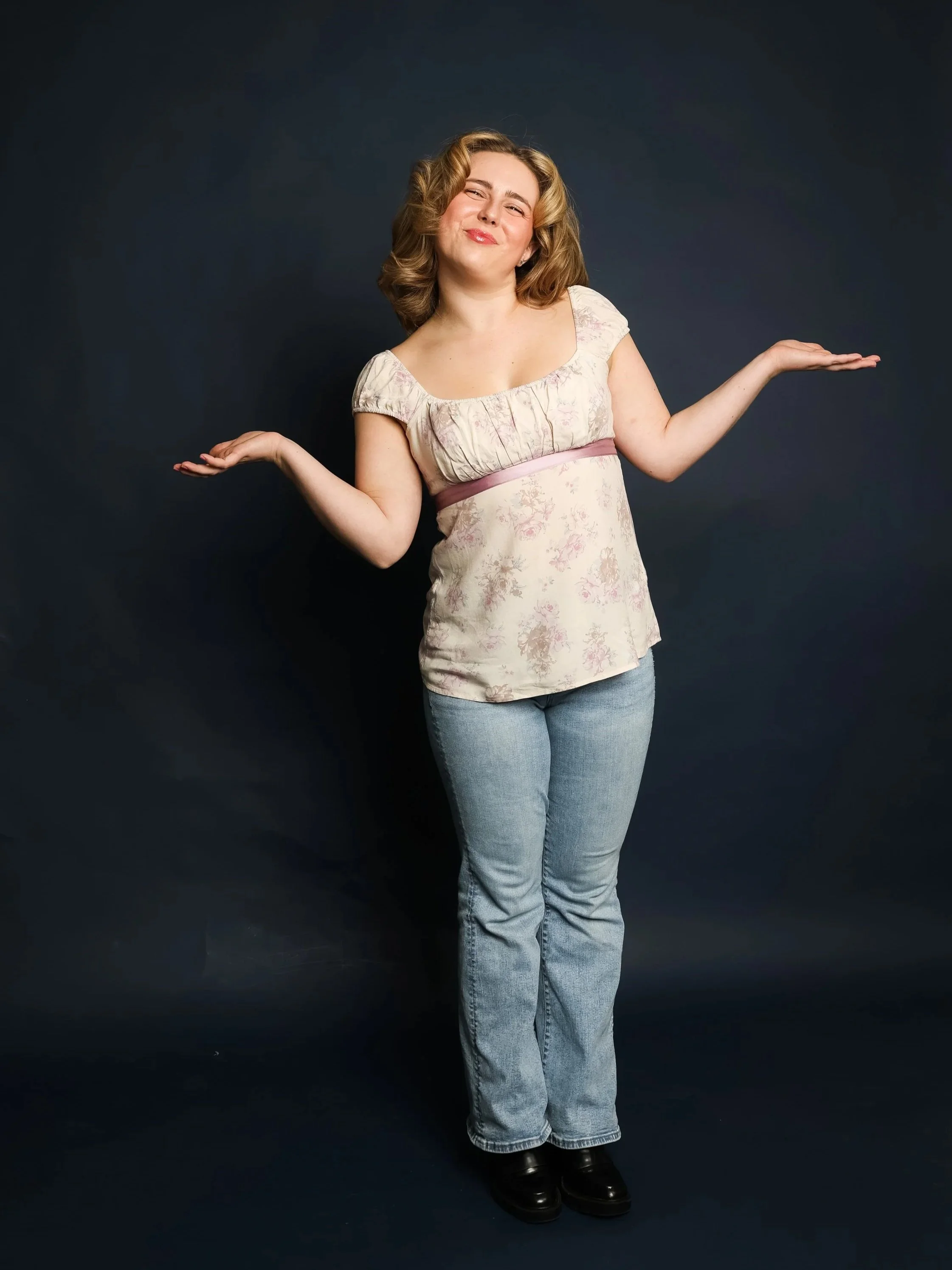 A woman with shoulder-length curly blonde hair, smiling and shrugging with her arms out to the sides against a dark background.