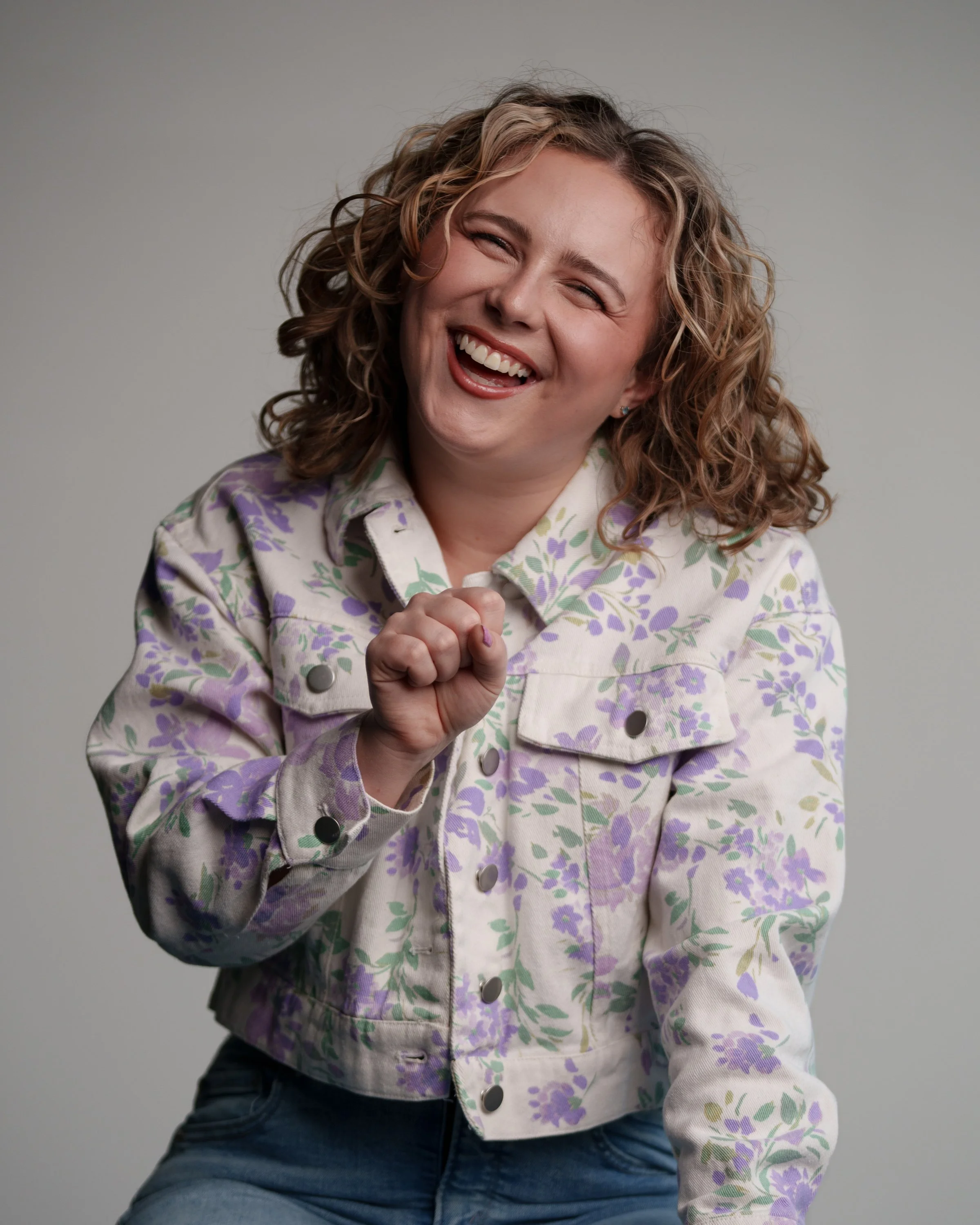 A woman with curly blonde hair laughing and smiling while making a fist with her right hand, wearing a floral patterned denim jacket and blue jeans.