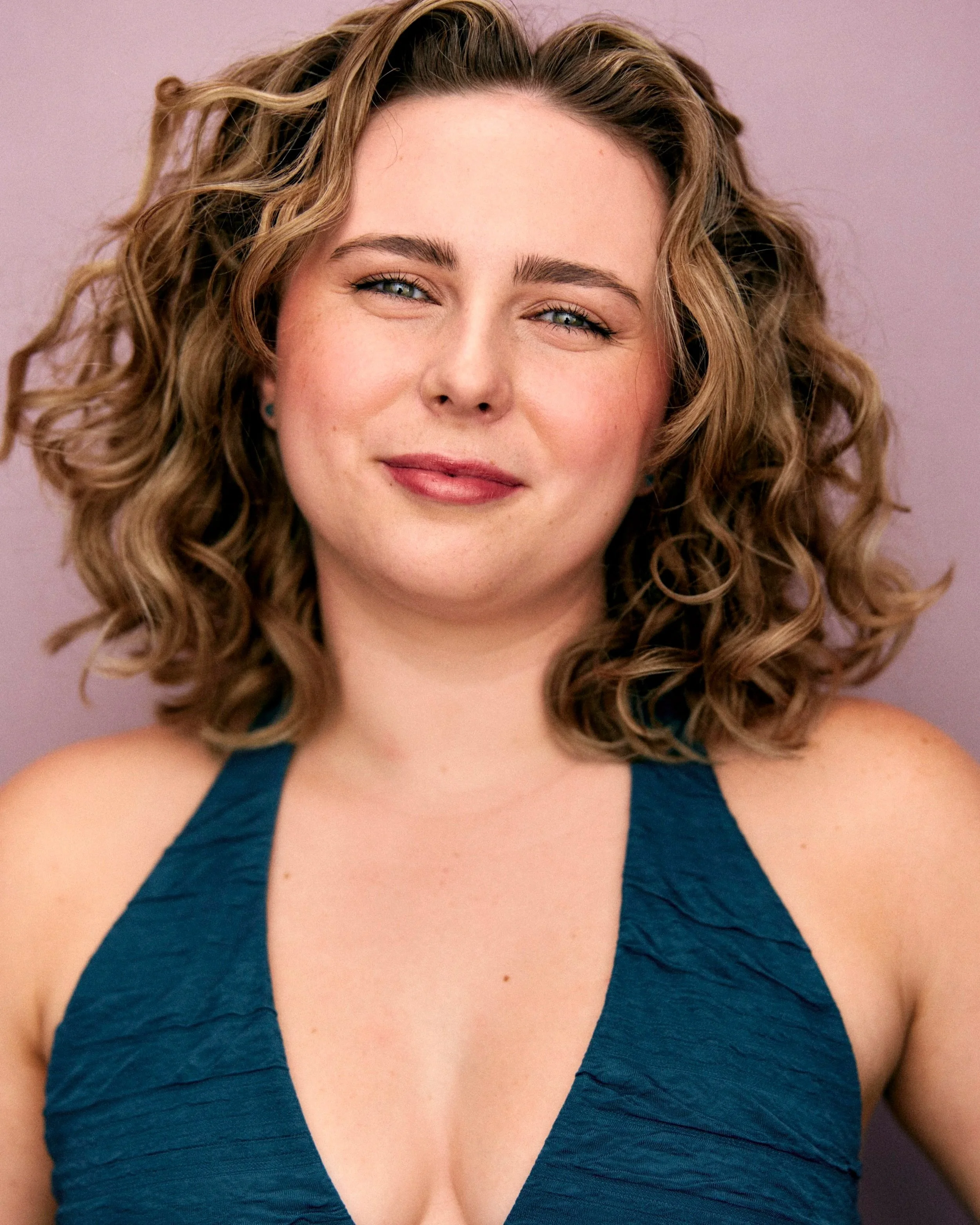 A woman with curly blonde hair, fair skin, and blue eyes smiling slightly, wearing a sleeveless dark teal top, against a light purple background.