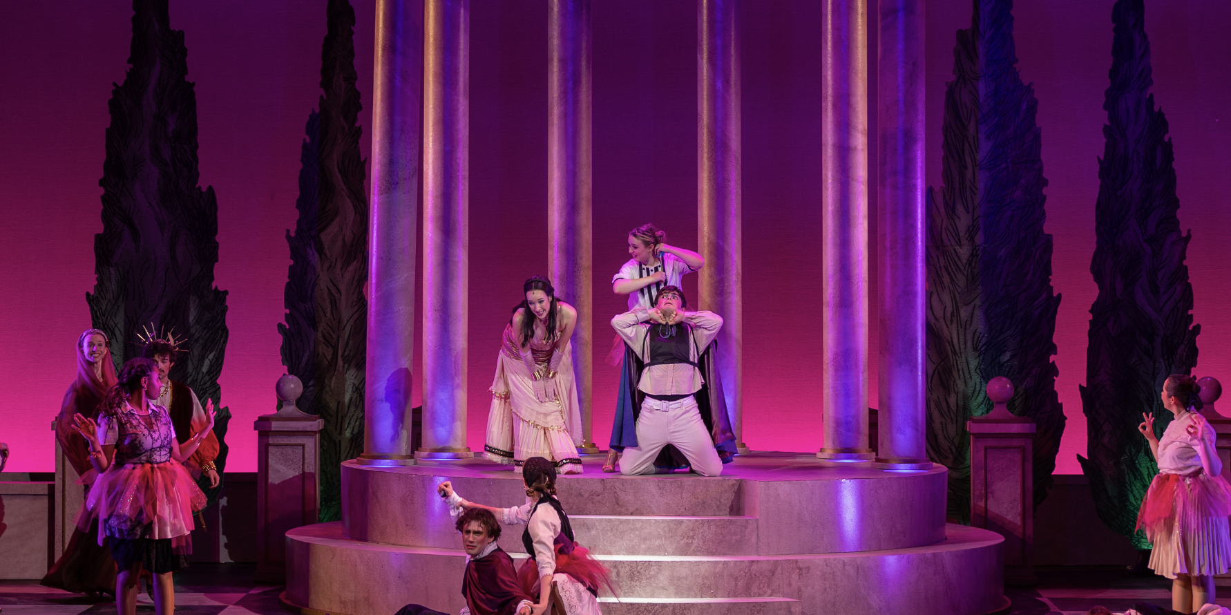 The stage scene from a theatrical production with actors dressed in colorful costumes, surrounded by tall columns and artistic backdrops, illuminated by purple and pink lighting.