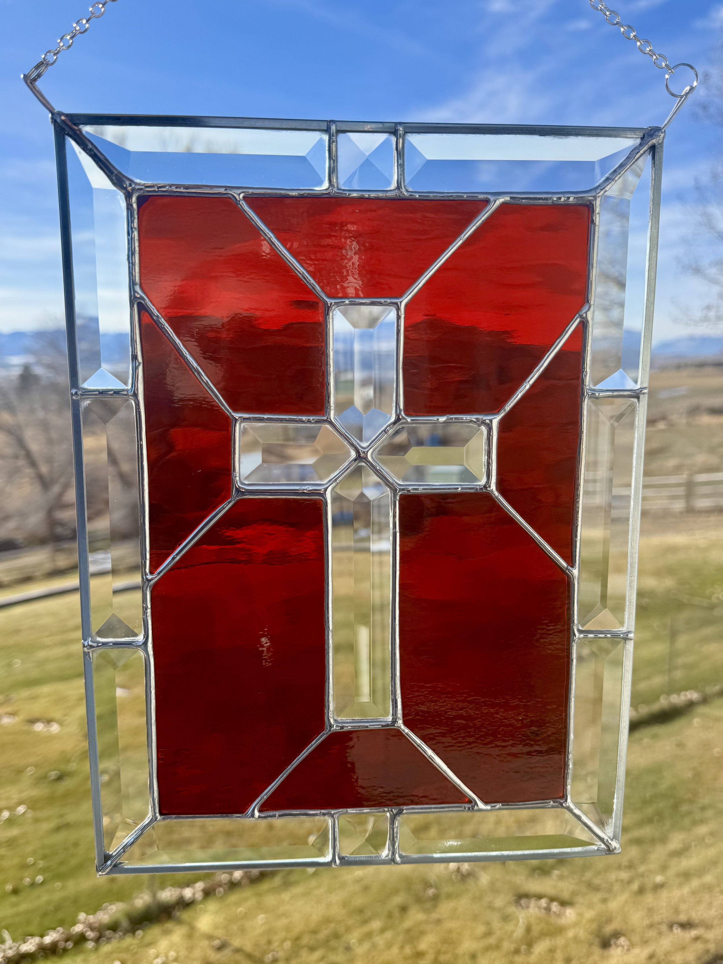 A decorative stained glass or glass art piece with geometric shapes, including a large red central panel, hanging outdoors against a background of a blue sky and landscape.