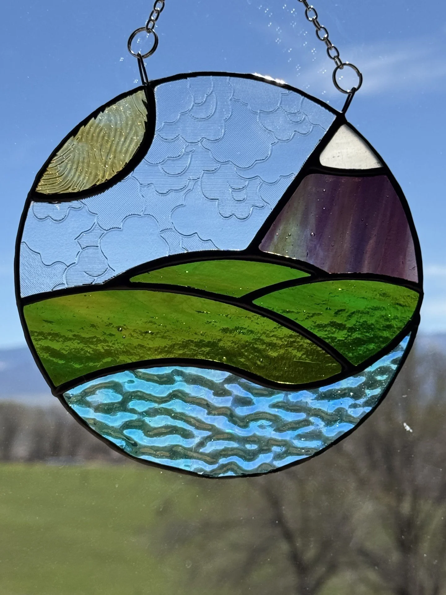 Colorful stained glass sun and landscape design hanging against a blue sky, with a grassy field below.