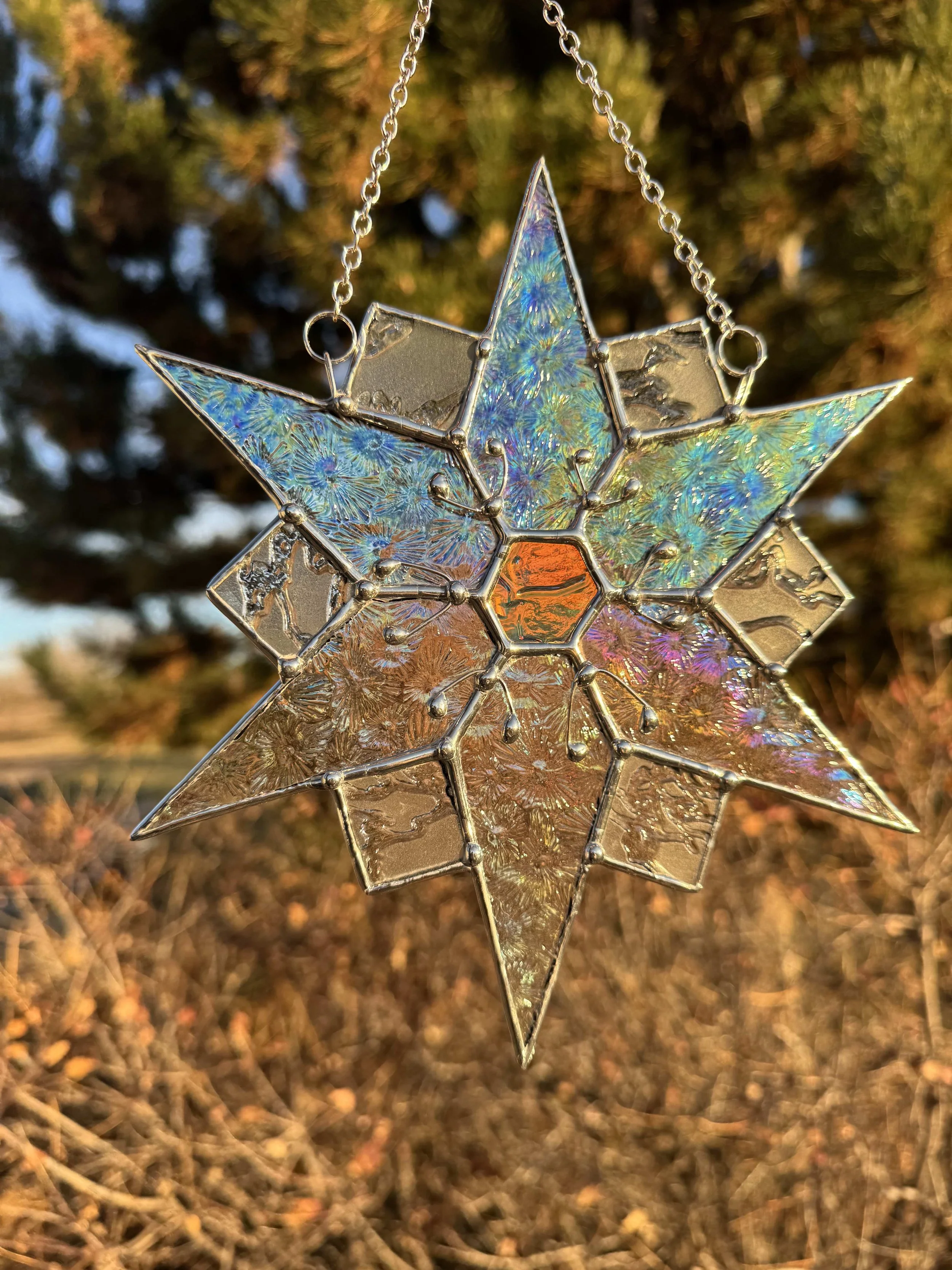 Close-up of a stained glass star ornament hanging outdoors with a blurred background of brown and green foliage.