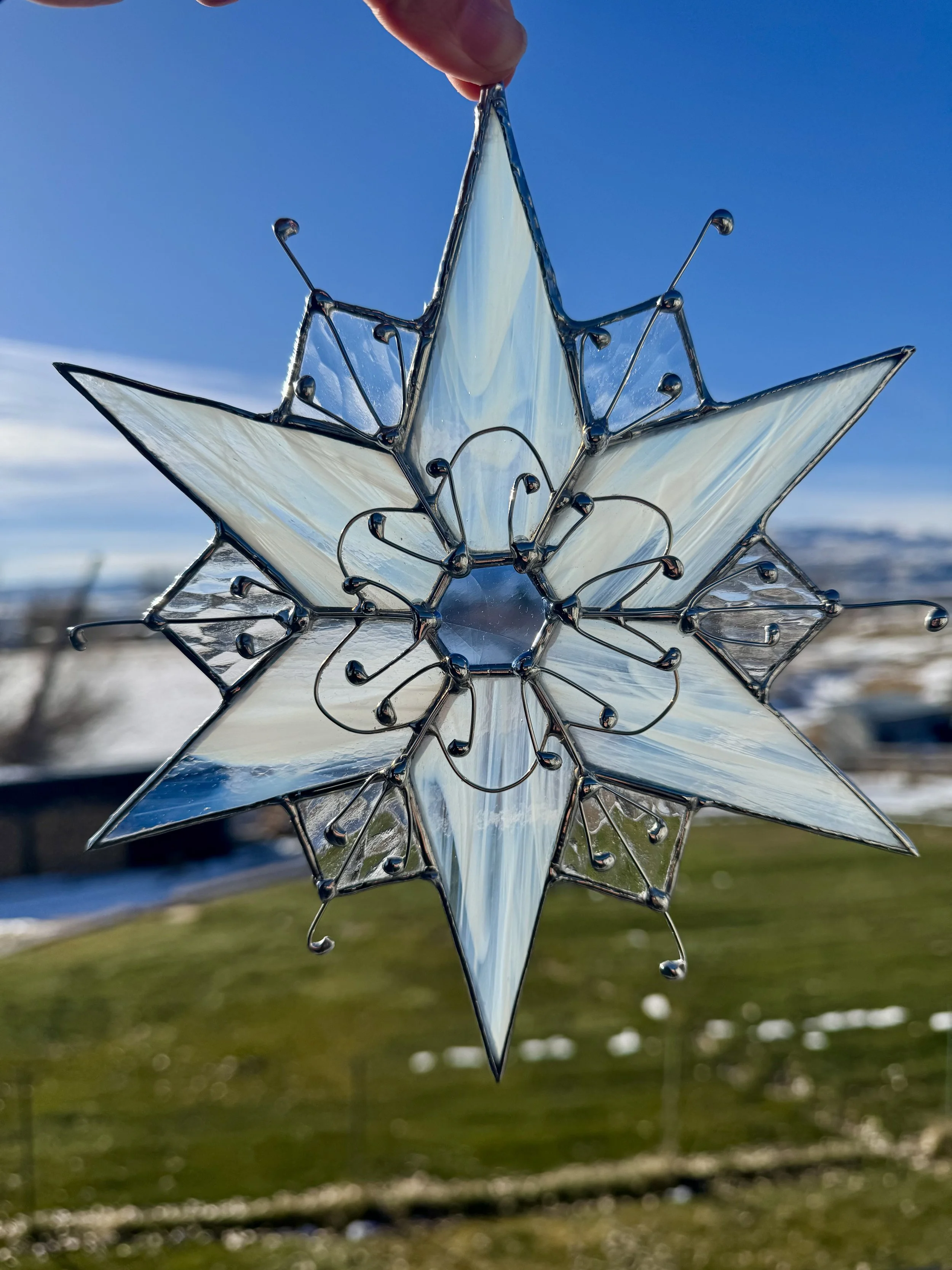 A close-up of a decorative snowflake ornament made of glass with wire detailing, held against a bright blue sky with a blurred landscape in the background.