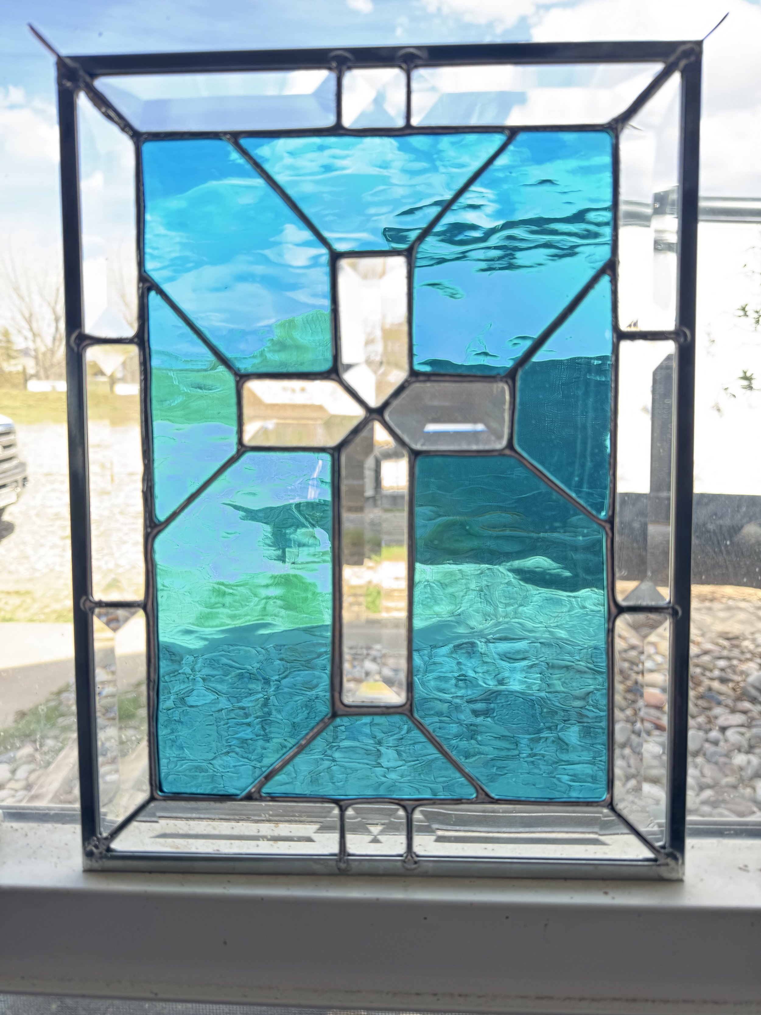 A stained glass window with a design resembling a bear, featuring shades of blue and black outlines, placed on a windowsill with an outdoor scene of trees, a house, and a cloudy sky in the background.