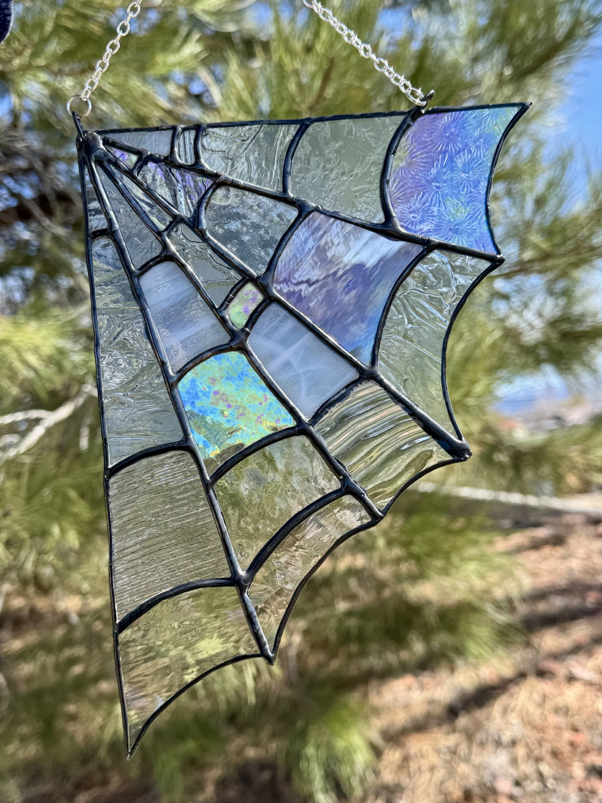Close-up view of a stained glass ornament in the shape of a spider web hanging outdoors, with a blurred background of trees and sky.