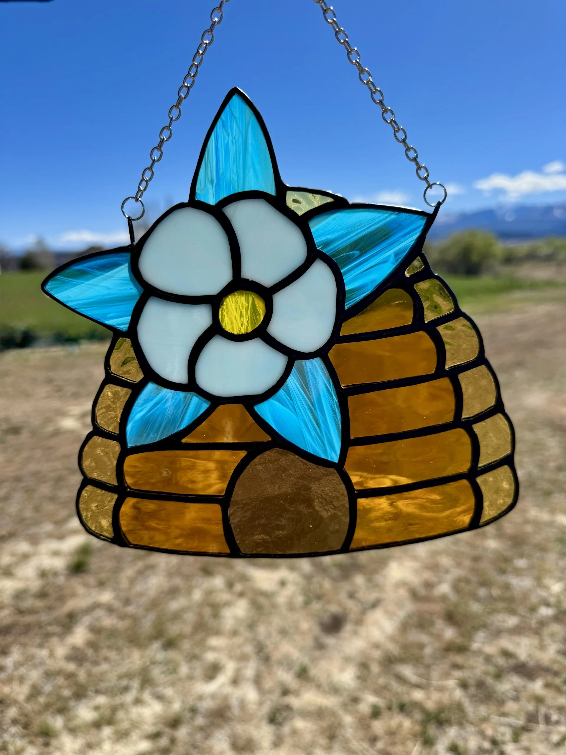 A stained glass sun catcher featuring a white flower with a yellow center and blue and amber petals, hanging on a chain against a clear blue sky with of a rural landscape in the background.